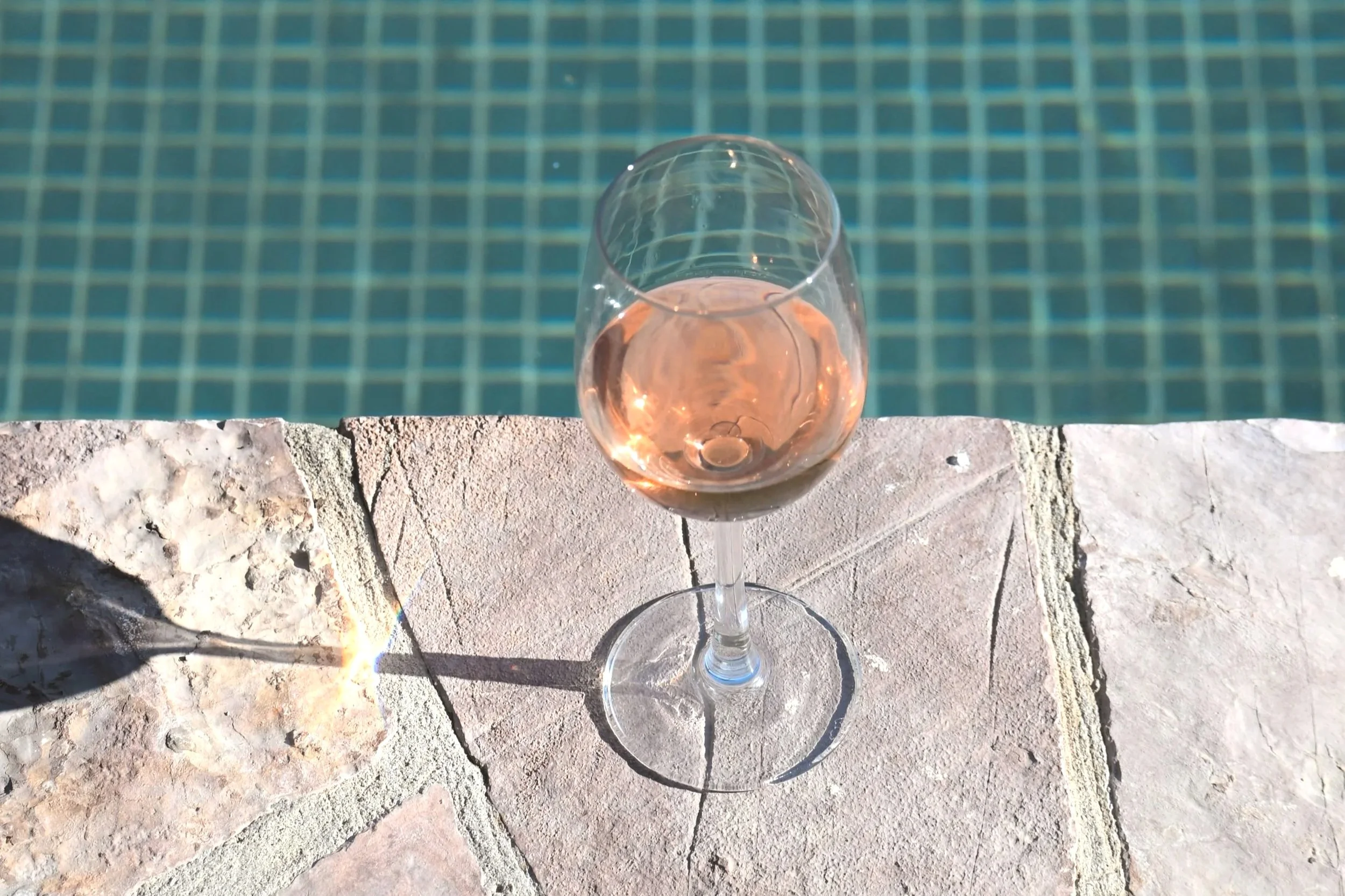 A glass of rosé wine on a stone surface next to a swimming pool.