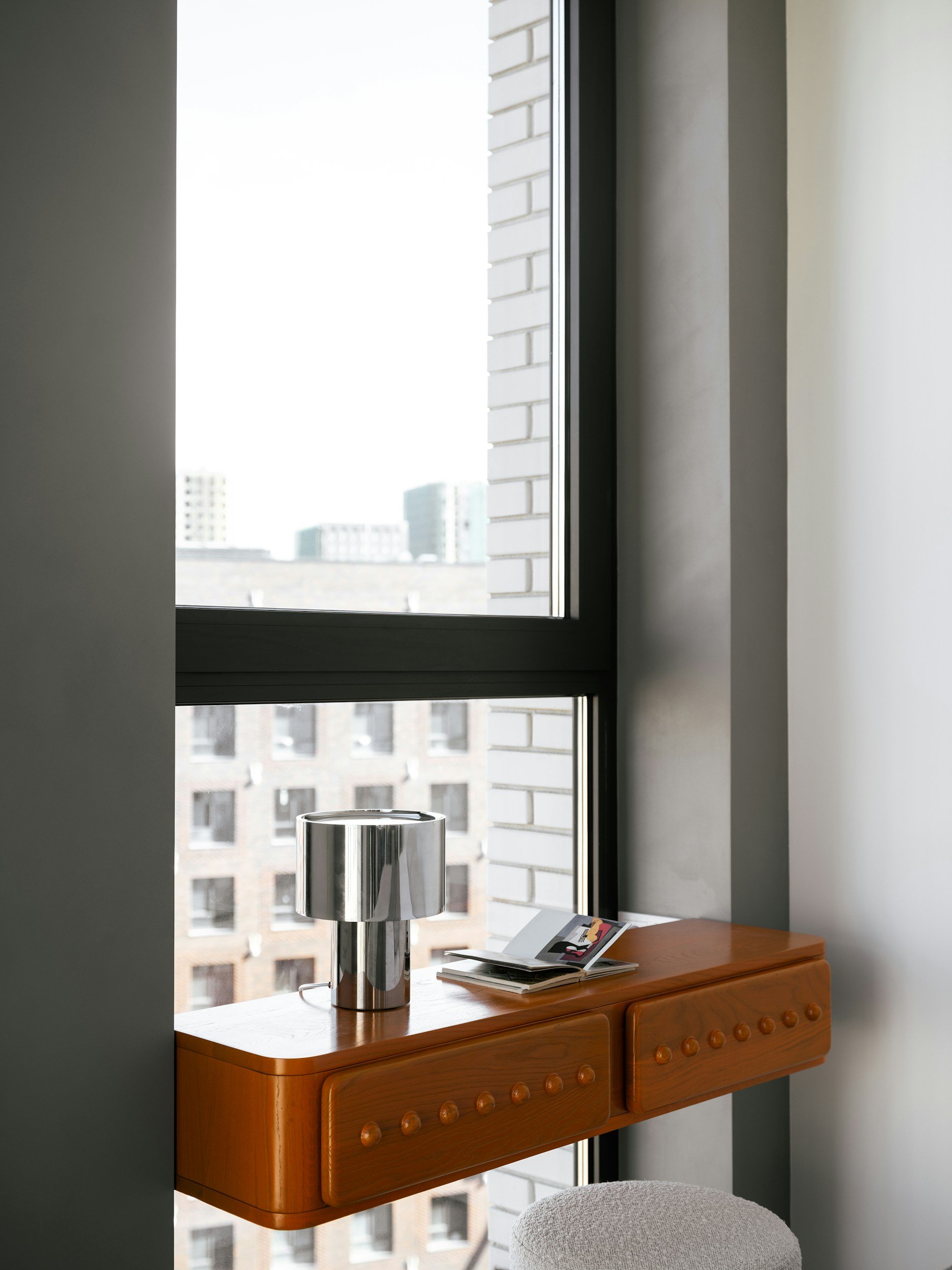 A window with a view of a cityscape, a wooden shelf with a metallic lamp and some papers, part of a chair in the foreground.