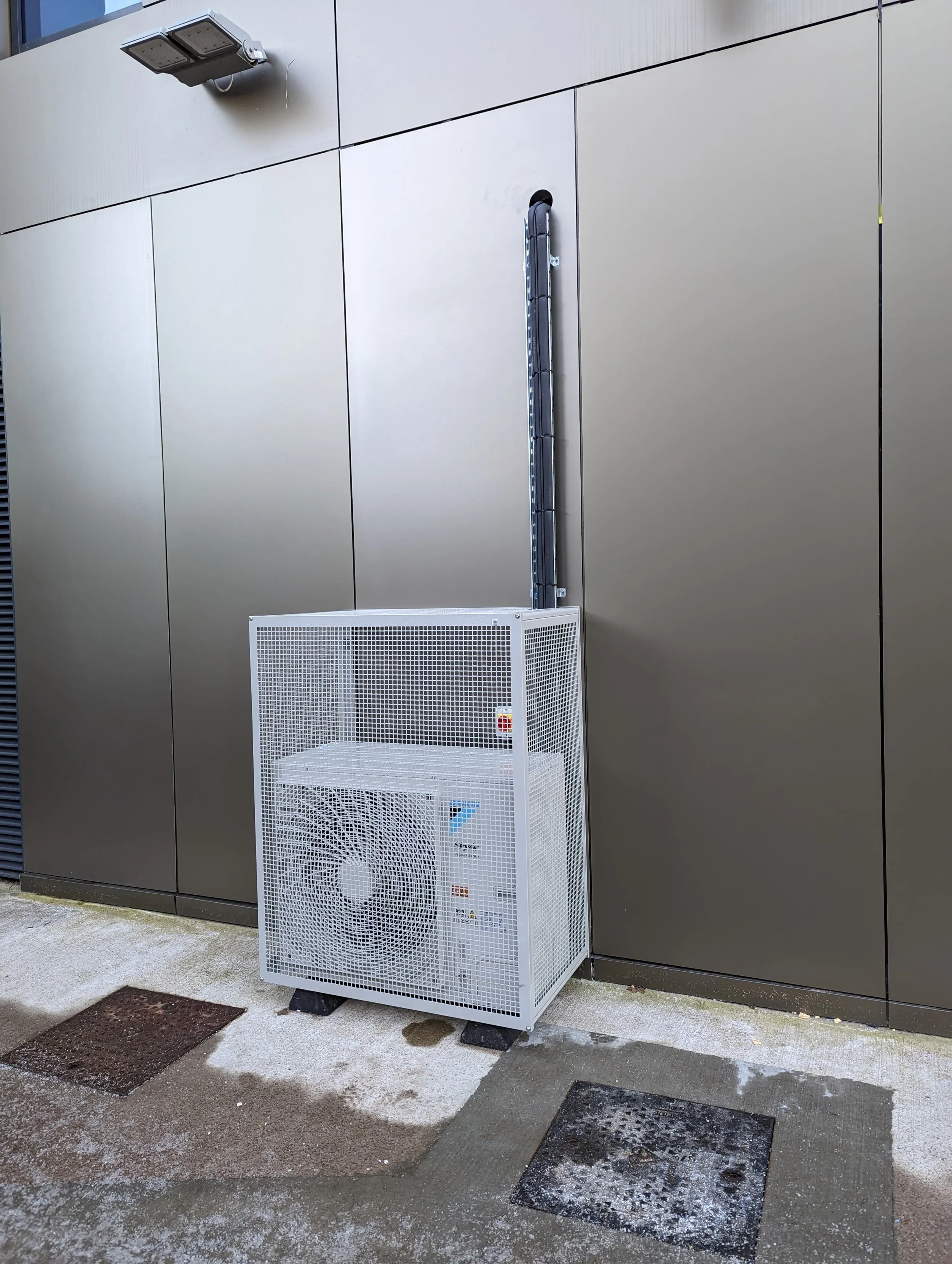 A white outdoor climate control unit with a protective metal mesh cover is positioned against a gray wall. The unit has a visible fan and control panel on its front. An industrial light fixture is mounted on the wall above.