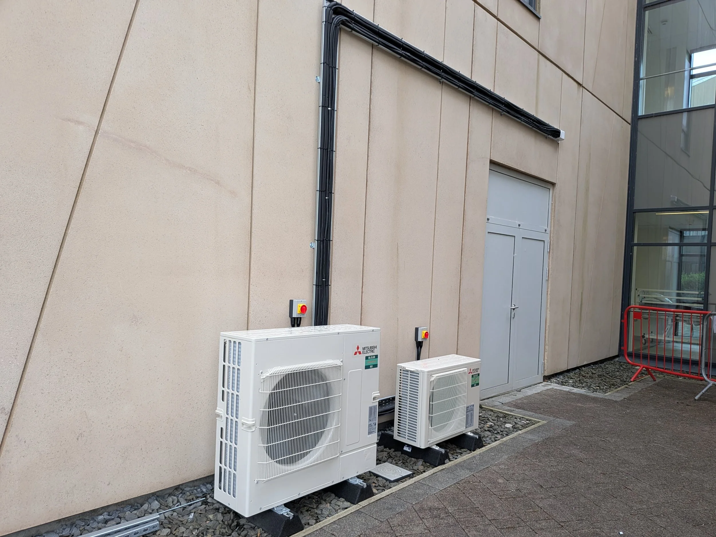 Outdoor scene showing two white air conditioning units mounted on black supports on a concrete surface beside a beige building with a gray door. Black electrical conduits run vertically along the building wall. There are two alert switches on the units and some metal barriers, along with a glass windowed section of the building.