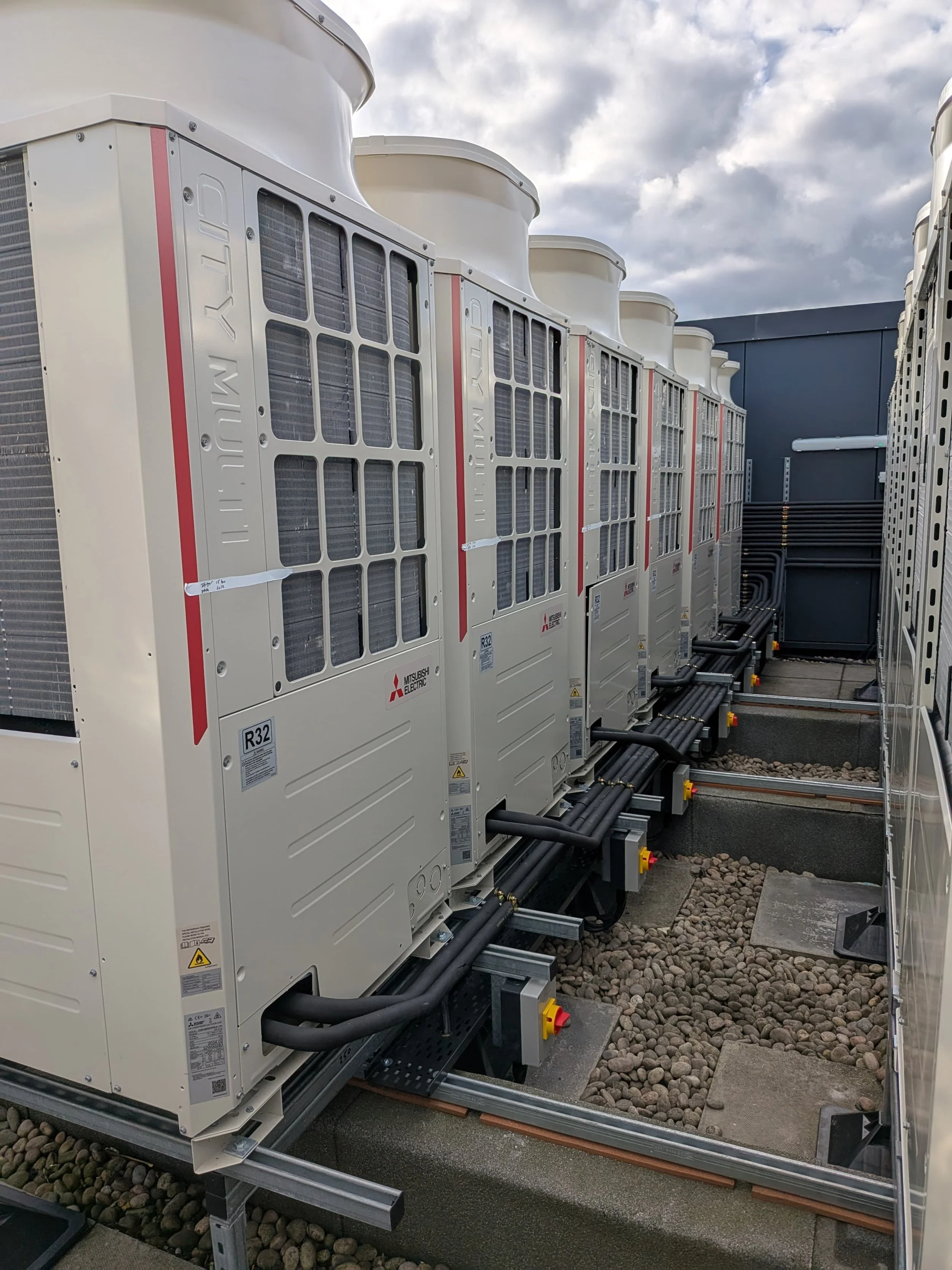 Multiple industrial HVAC units installed outdoors on a rooftop, with electrical and piping connections, under a cloudy sky.