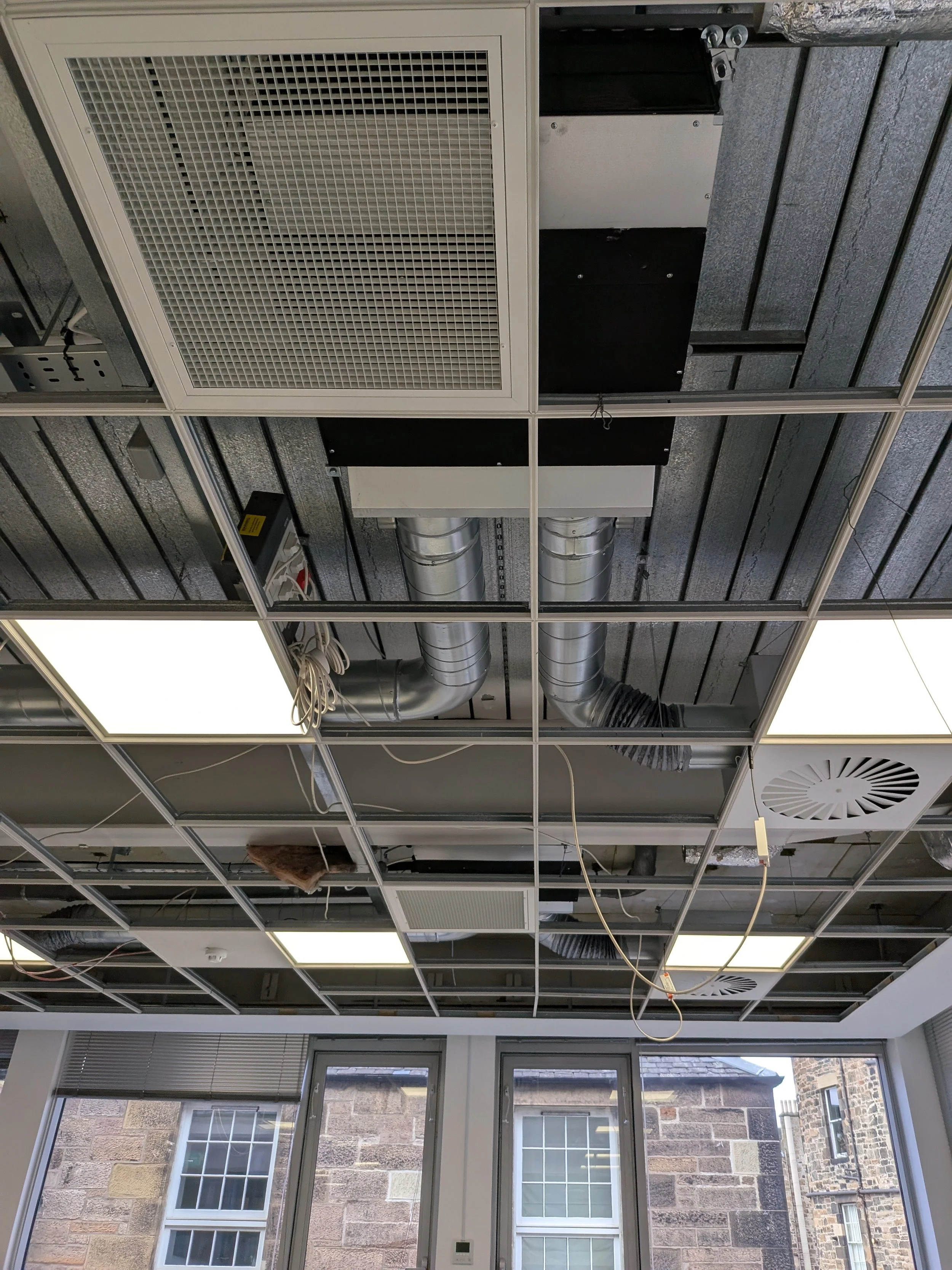 Ceiling with exposed ductwork, ventilation, and lighting fixtures, with large windows showing a stone building outside.