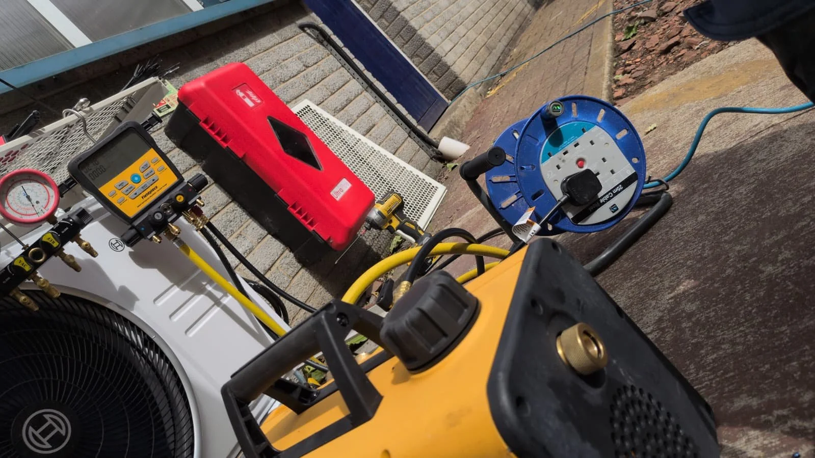 Outdoor scene with HVAC equipment and electrical tools, including a yellow generator, a red component, a blue reel with extension cord, and a digital gauge, placed on a concrete surface near a building.