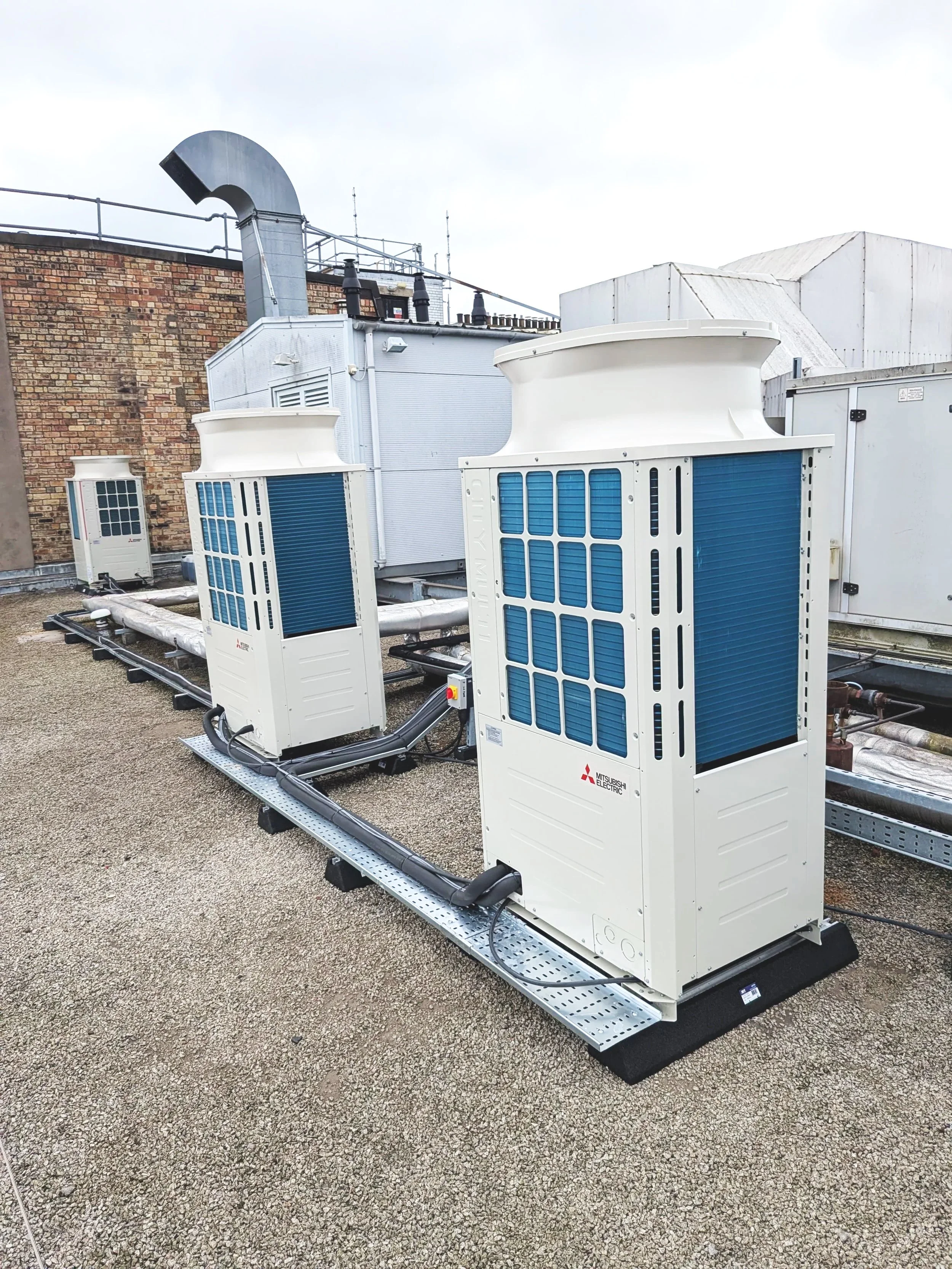 Rooftop HVAC units with blue vents, connected by black pipes, on a flat rooftop with gravel surface, background features a brick wall and building structures.