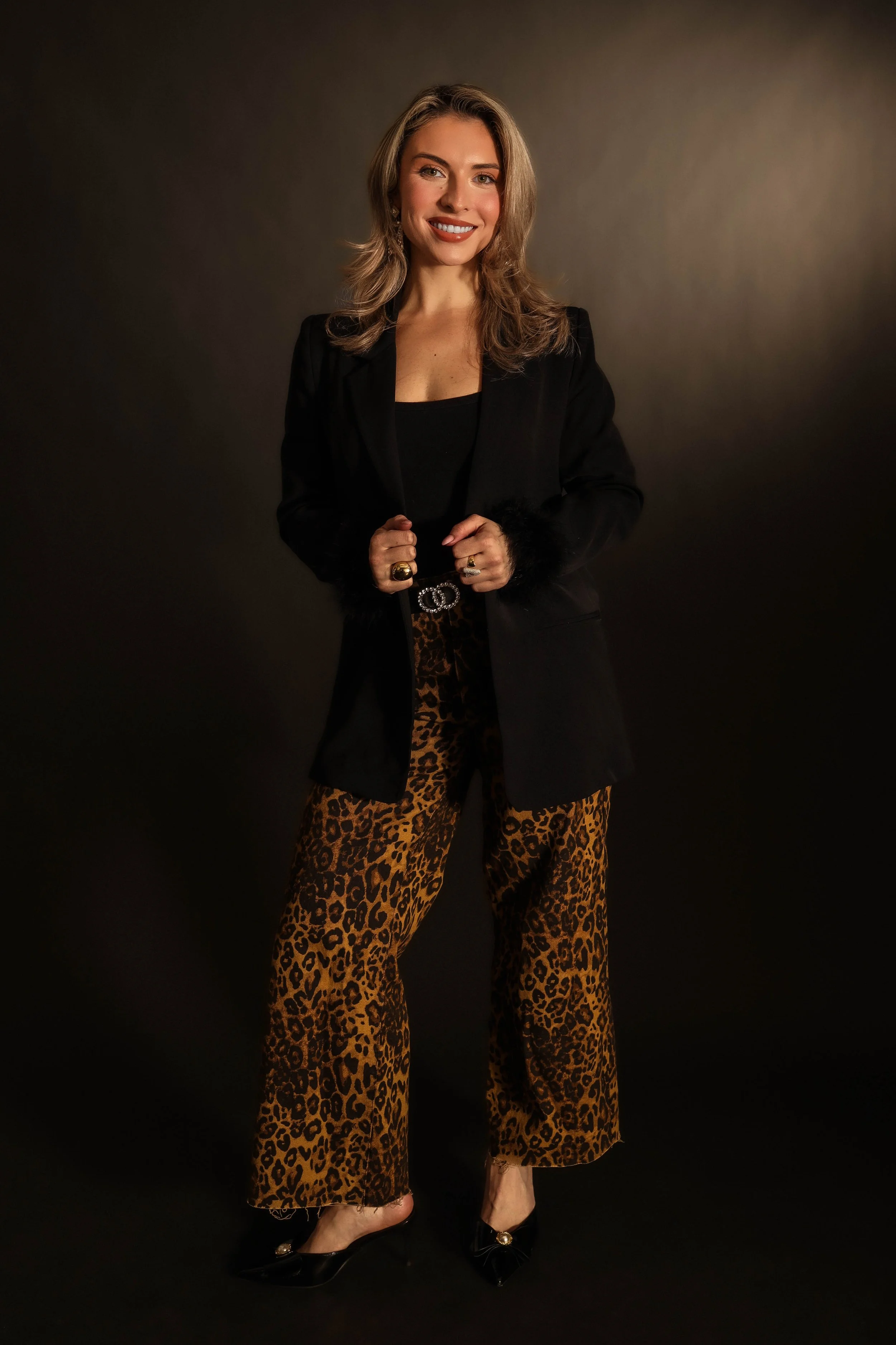 A woman with shoulder-length blonde hair and a big smile, standing against a dark gradient background. She is wearing a black blazer, leopard print wide-leg pants, black high heels, and has rings on her fingers.