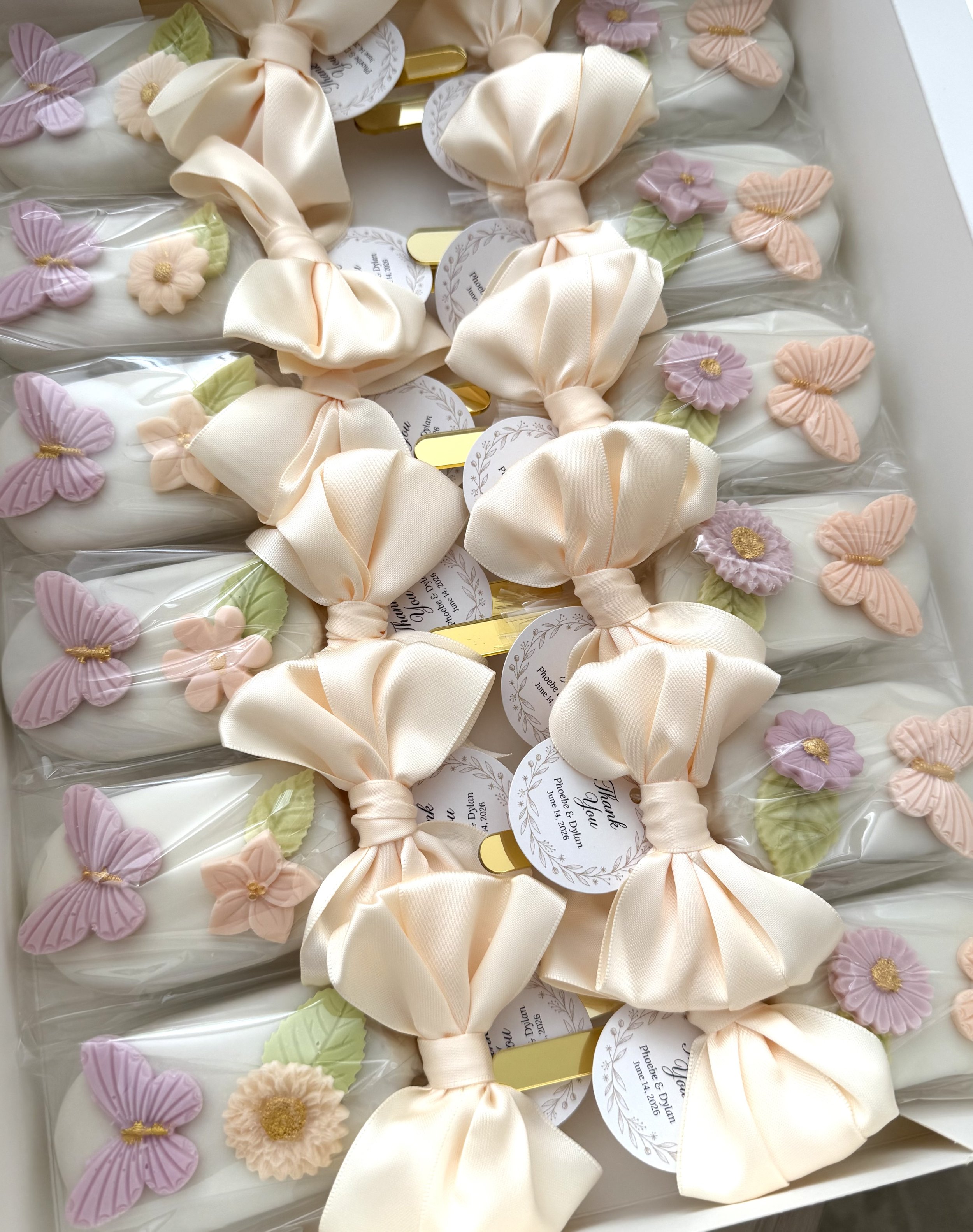 Decorative cakepops wrapped with beige satin ribbons, adorned with small fabric flowers and butterflies in purple and peach colors, arranged in a box for wedding reception.