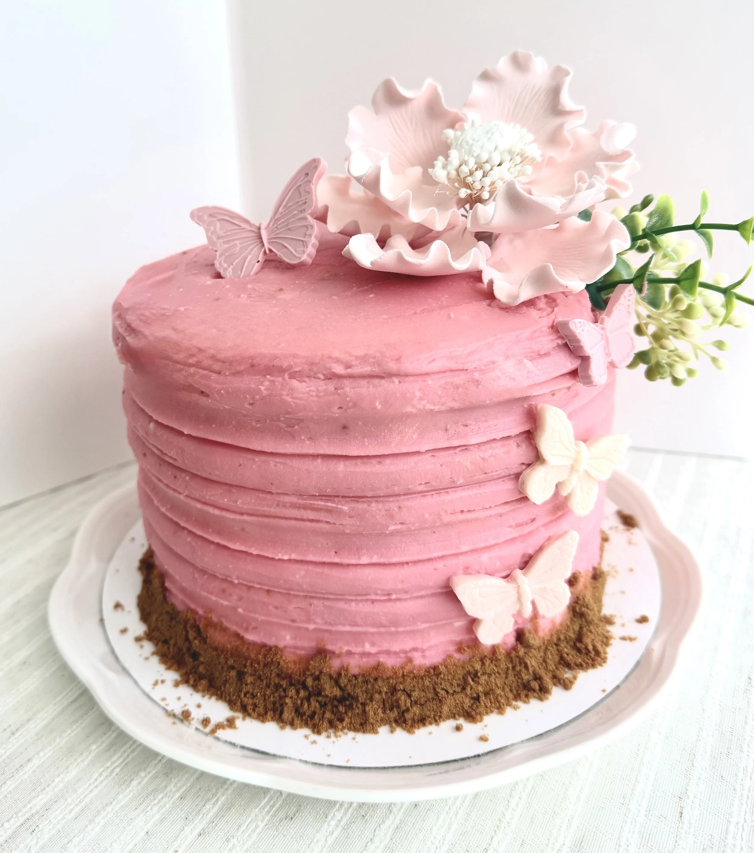 Pink layered cake with chocolate crumbs on the bottom, decorated with white and pink butterflies, a pink flower, and green foliage on top.