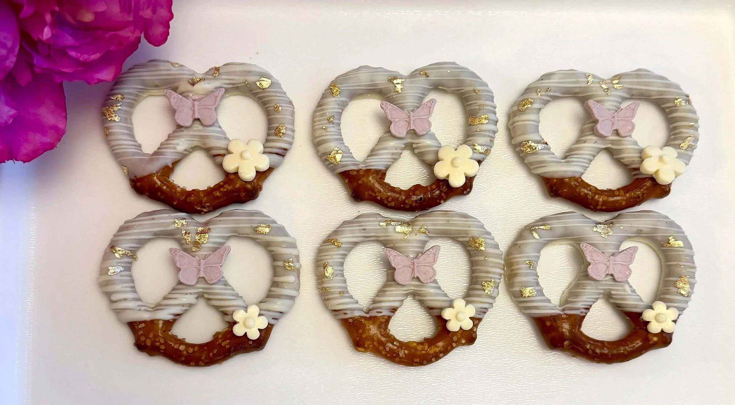 A row of six pretzels decorated with white and pink icing, each topped with a small pink butterfly and a white flower, and sprinkled with gold leaf.