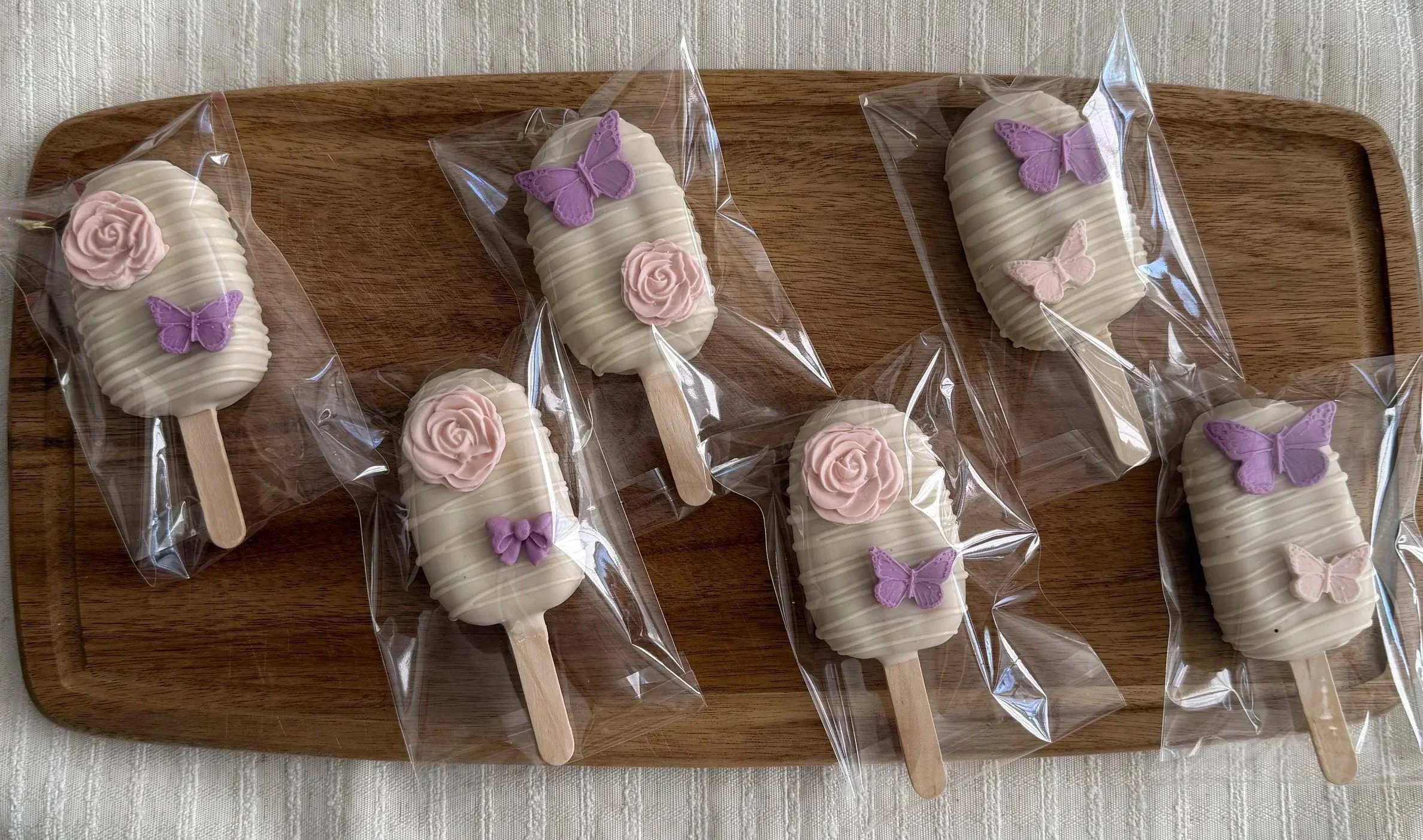Treats wrapped in clear plastic, decorated with pink roses and purple butterflies, placed on a wooden tray.
