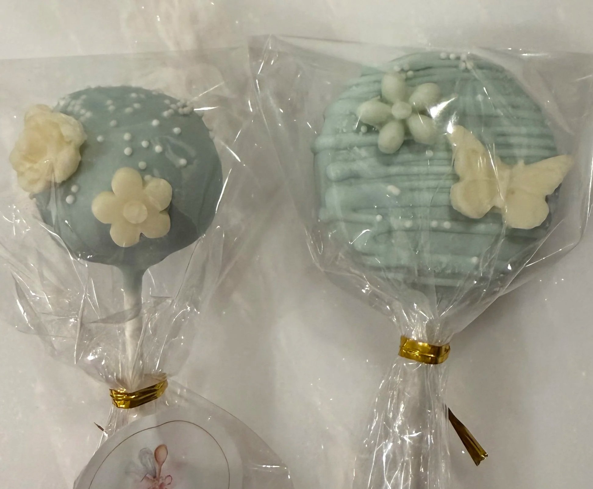 Two wrapped lollipops with white flower decorations inside clear plastic wrapping.