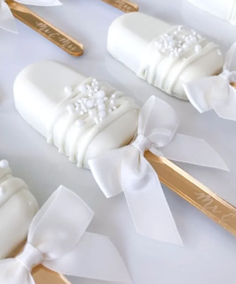 White chocolate-covered Oreo bars decorated with white pearls, each tied with a white ribbon and attached to a wooden stick.