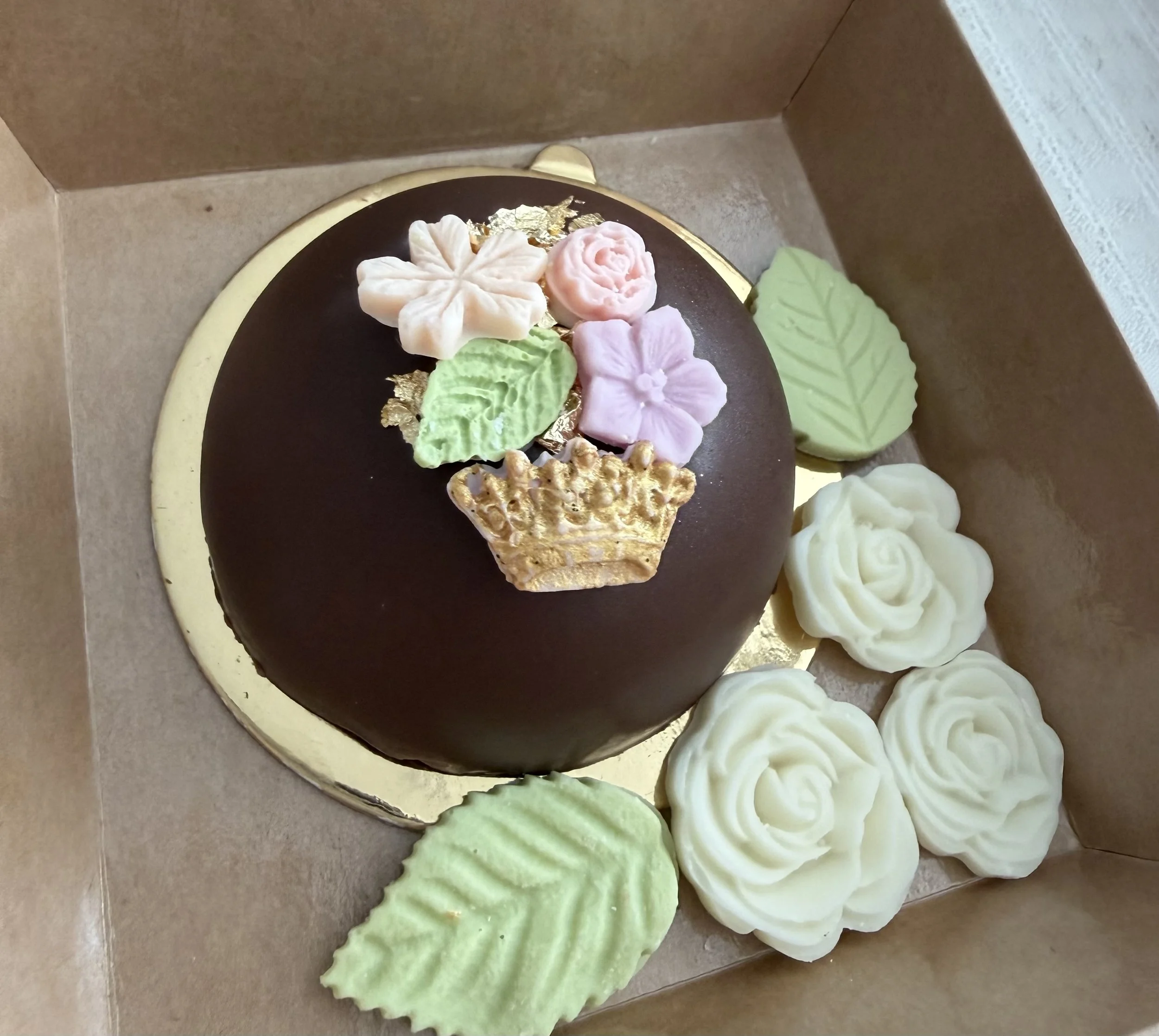 Dubai style pistachio dome decorated with flower and leaf-shaped chocolate, placed inside a beige box with additional white and green icing flowers and leaves around it.