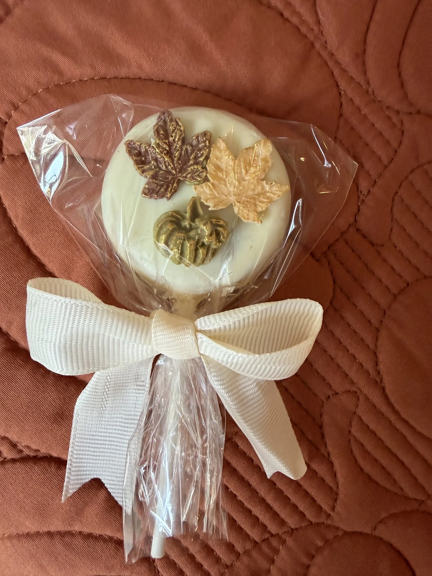 A decorated white chocolate cake pop with a white ribbon tied in a bow, featuring gold and brown maple leaf and acorn decorations, wrapped in clear plastic, placed on a textured quilted fabric surface.