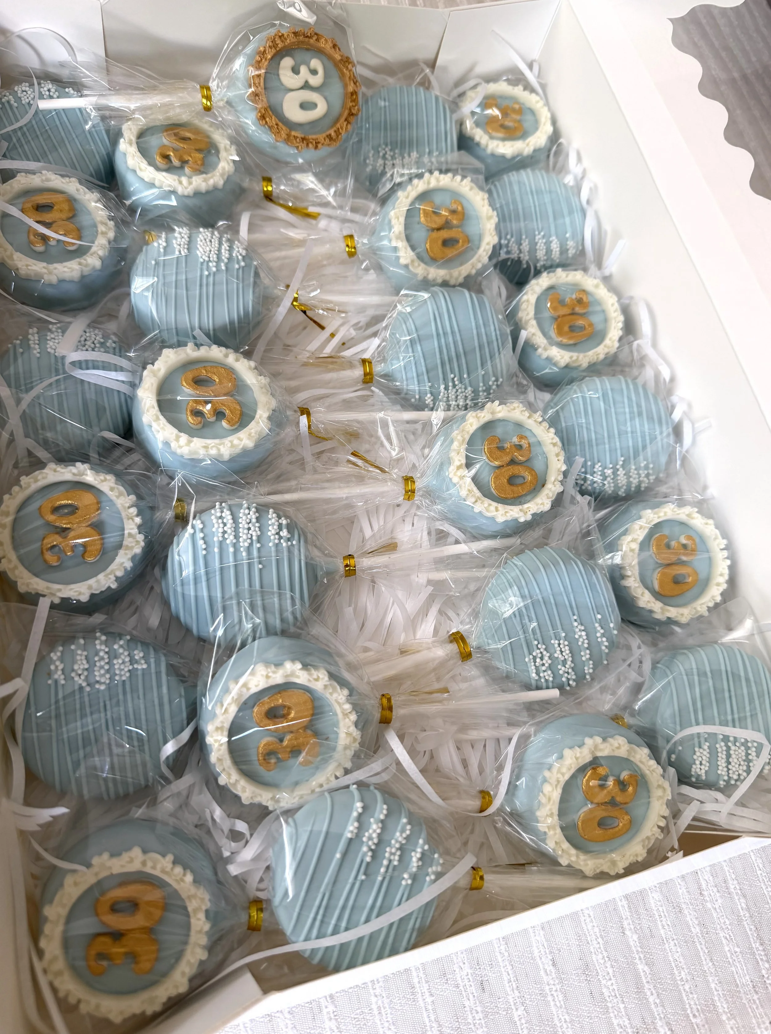 A white box filled with decorated cakepops wrapped in plastic. The cakepops are light blue with white and gold details, featuring numbers 30 for thirties birthday.