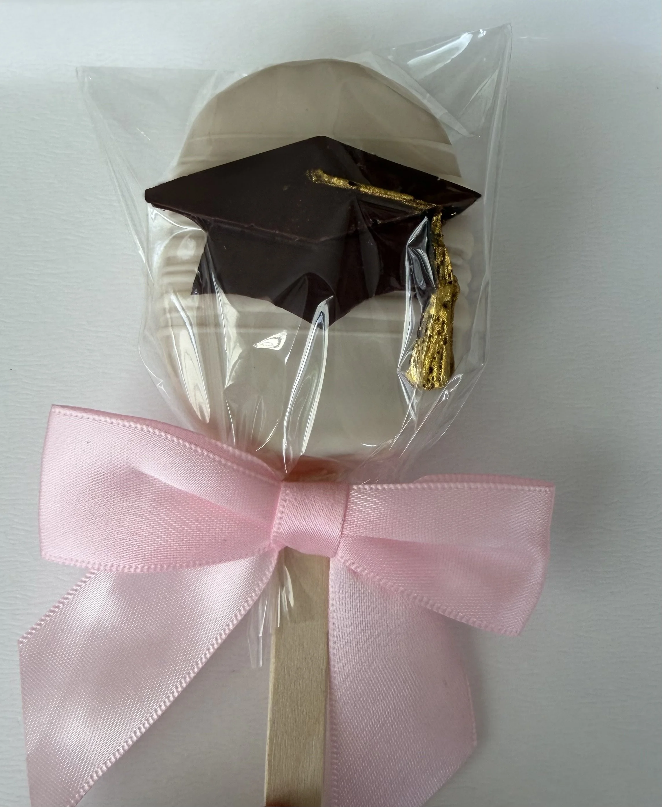 A small decorative graduation cap with a gold tassel, wrapped in clear plastic, attached to a pink ribbon bow, placed on a white surface.