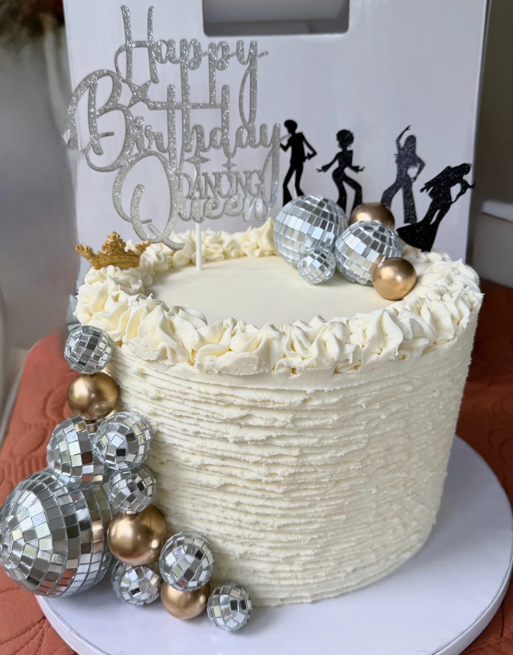 A birthday cake decorated with white cream cheese, metallic and gold disco ornaments, and black silhouette cake toppers of people dancing. A silver glittery "Happy Birthday Dancing Queen" as cake topper.