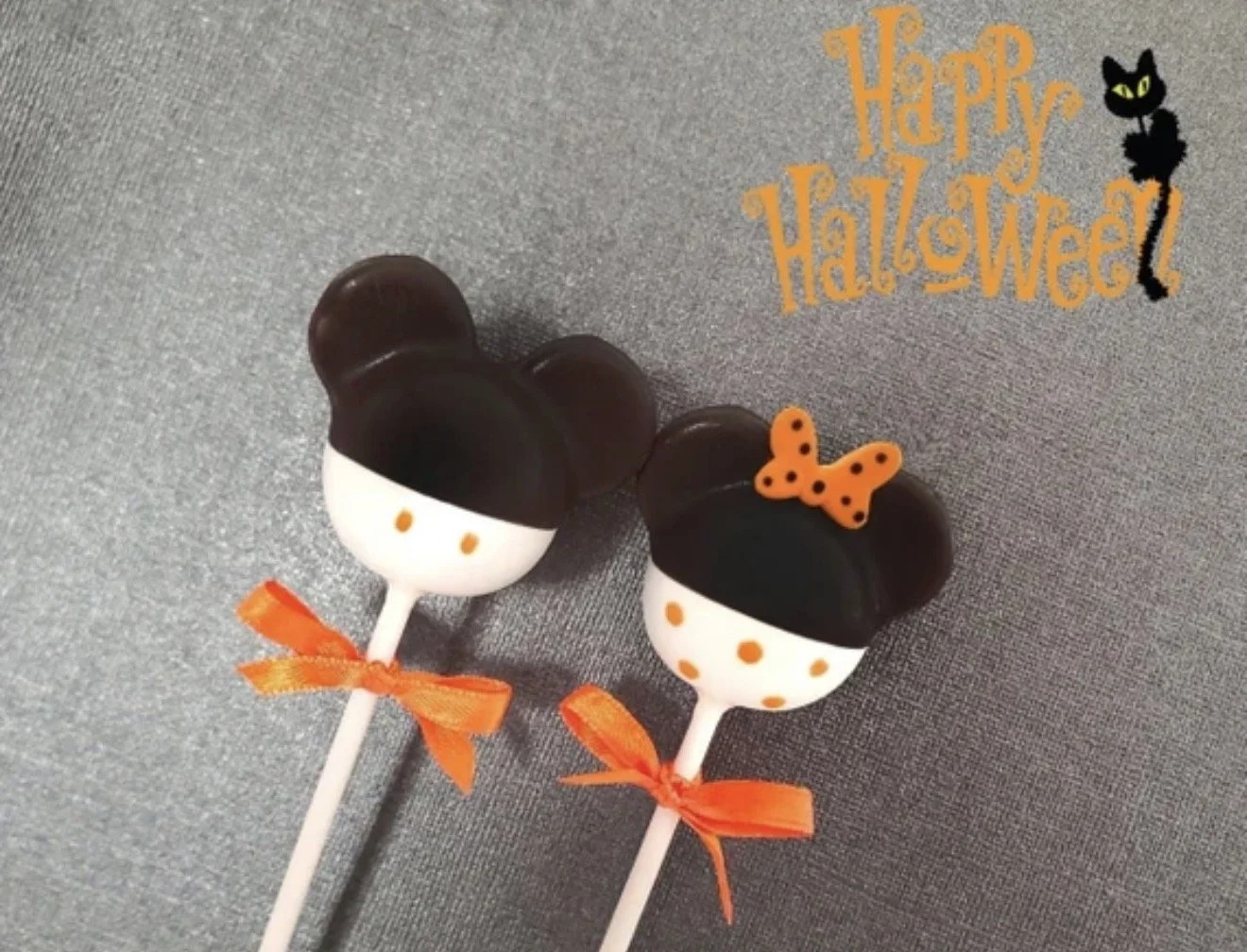Halloween-themed cake pops with Mickey Mouse and Minnie Mouse ears, decorated with orange bows and polka dots, on sticks with orange ribbons, a Halloween party treats.