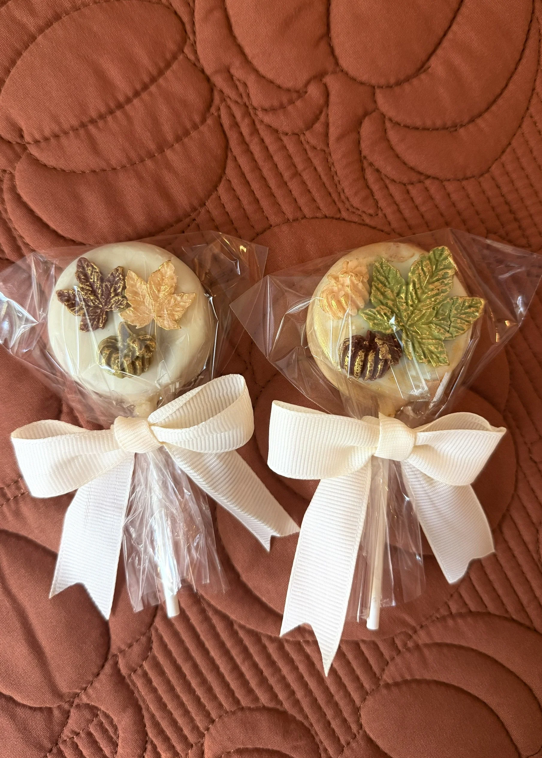 Two decorated cake pops featuring gold and brown maple leaf and acorn decorations, in clear plastic with white bows, placed on a quilted orange table runner.