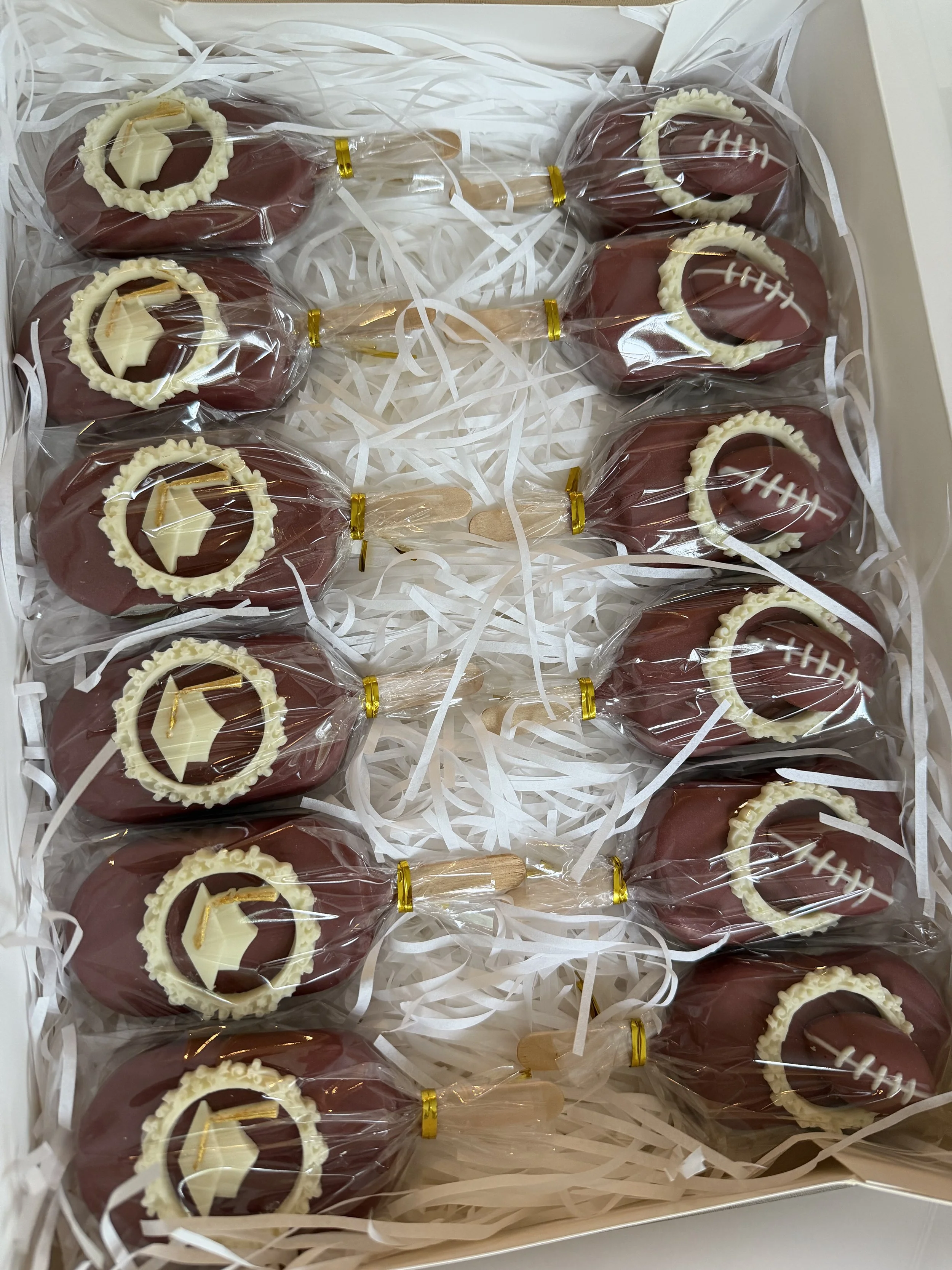 Box of chocolate cake pops decorated with white and dark chocolate, some with football stitching details, wrapped in clear plastic and arranged on shredded paper in a white box.
