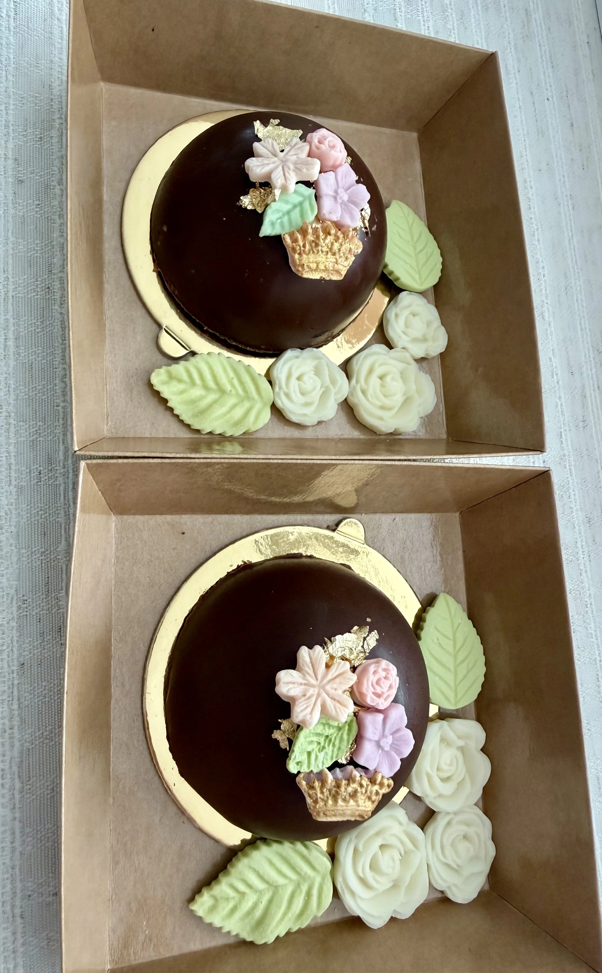 Dubai style pistachio dome decorated with flower and leaf-shaped chocolate, placed inside a beige box with additional white and green icing flowers and leaves around it.