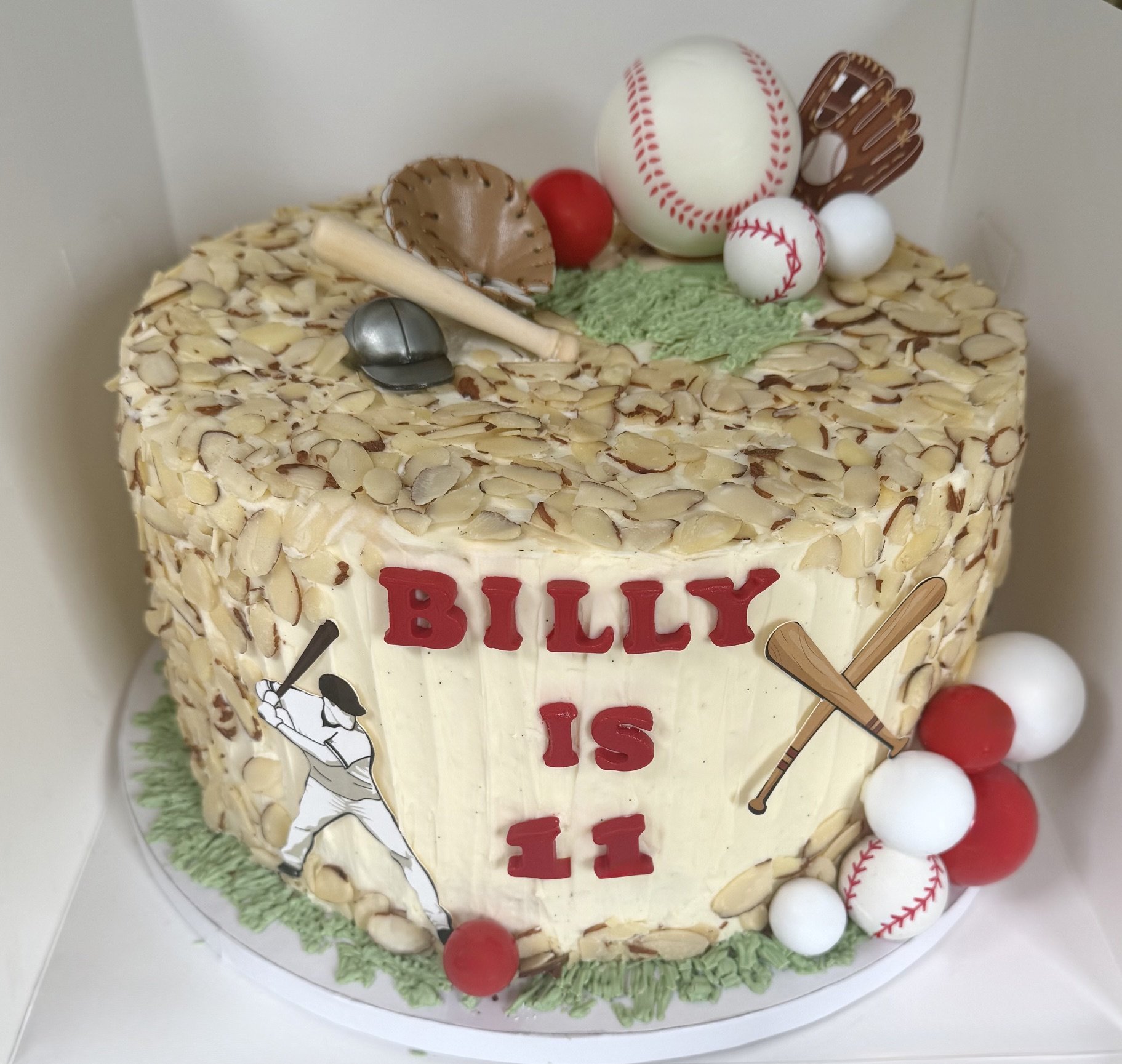 A baseball-themed birthday cake with decorative baseballs, a miniature baseball glove, and a sports-themed topper, surrounded by red and white balls, with green icing resembling grass on the cake.