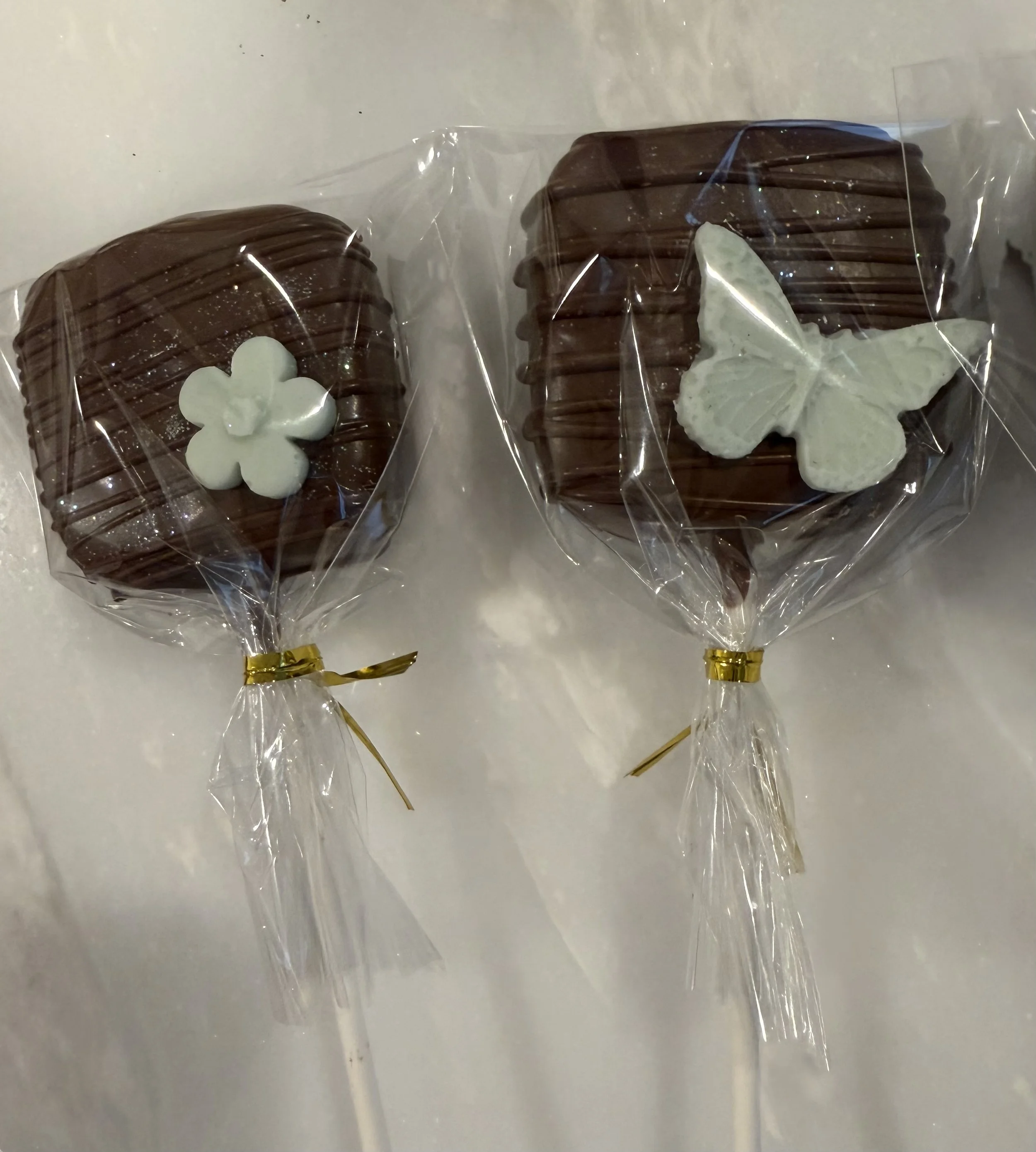 Two chocolate decorated treats wrapped in clear plastic with white flower and butterfly decorations, tied with gold ribbons.