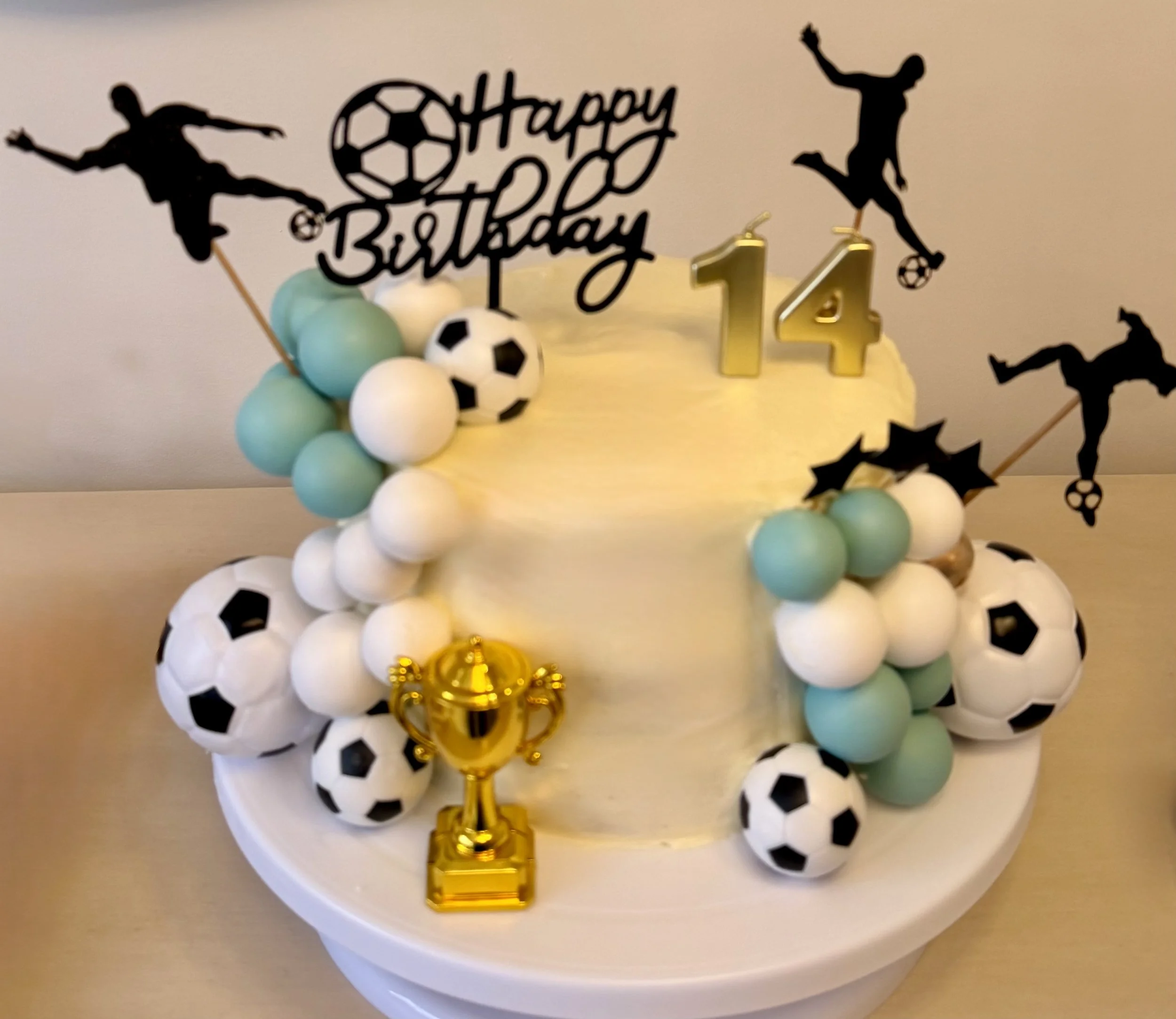 A birthday cake decorated with blue and white balls, black and white soccer ball decorations, a gold birthday candle shaped like the number 14, and a cake topper reading 'Happy Birthday' with silhouettes of soccer players.