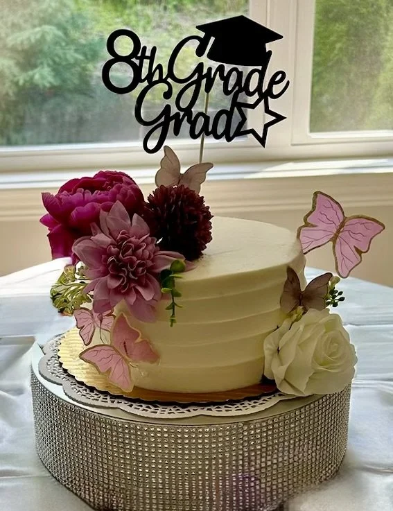 A graduation cake decorated with pink and white flowers, butterfly toppers, and a "8th Grade Graduation" sign with a graduation cap, placed on a table near a window.