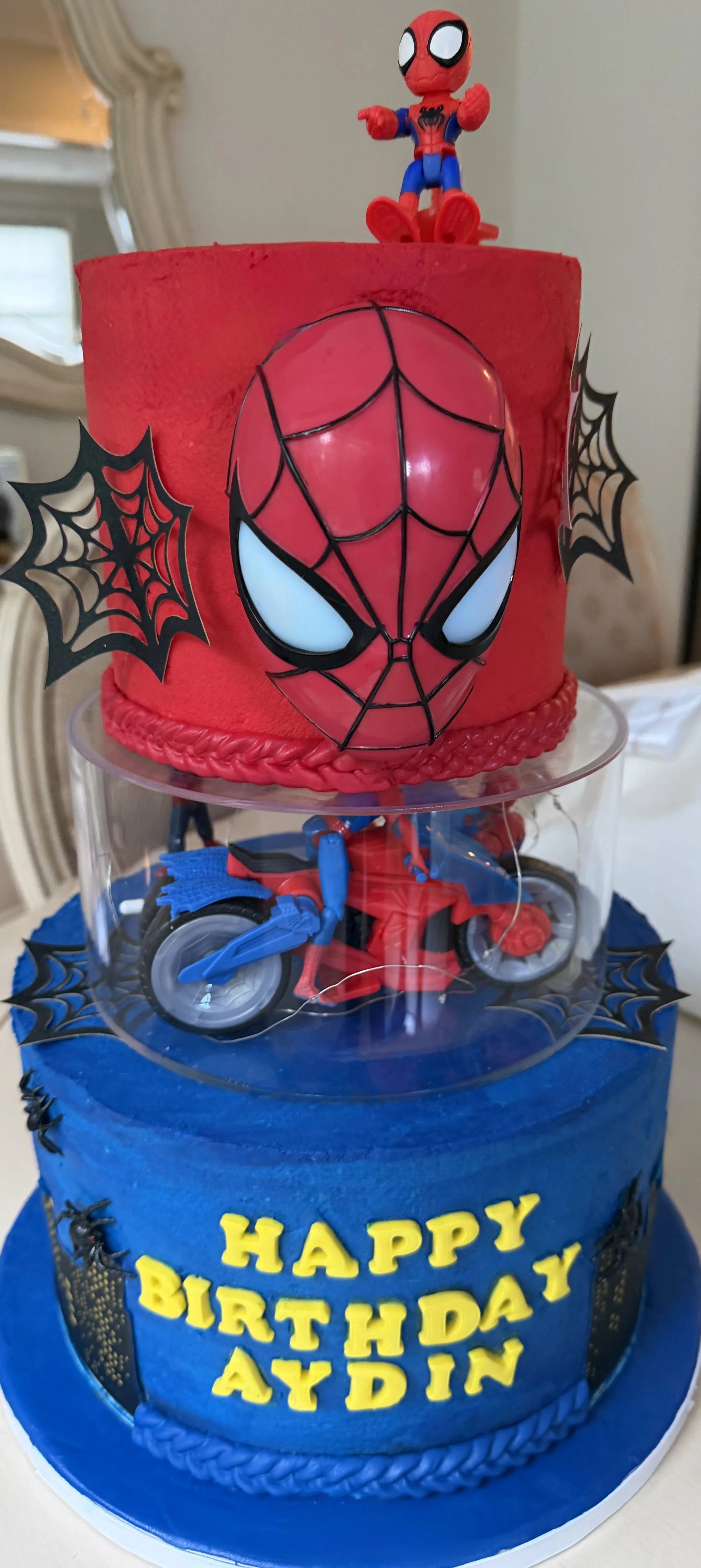Three tier Spider-Man themed birthday cake with a Spiderman face design, web decorations, and figurines of Spider-Man and a motorcycle, with a birthday letters "Happy Birthday Aydin" in yellow fondant.