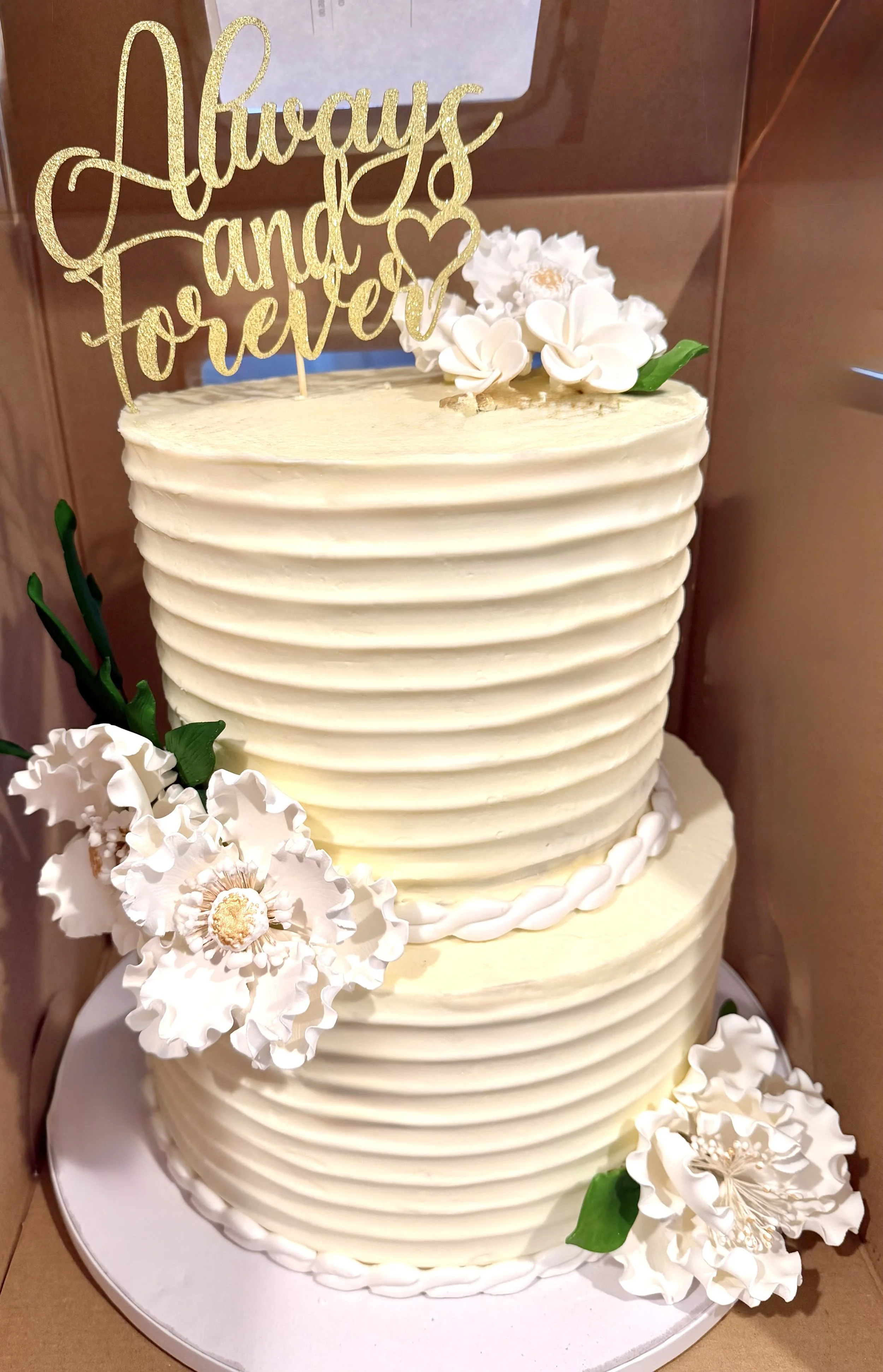 A multi-tiered wedding cake decorated with white edible flowers and green leaves, topped with a gold glittery cake topper that reads 'Always and Forever'.