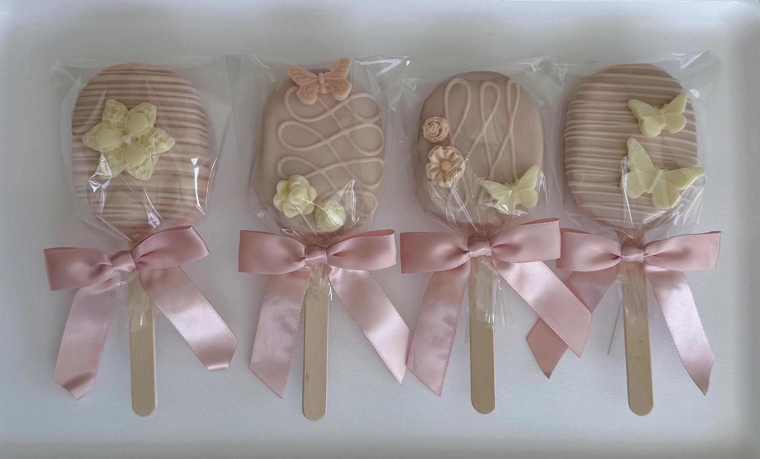 Four pastel-colored decorated cookies wrapped in plastic and tied with pink ribbons, each attached to a wooden popsicle stick.