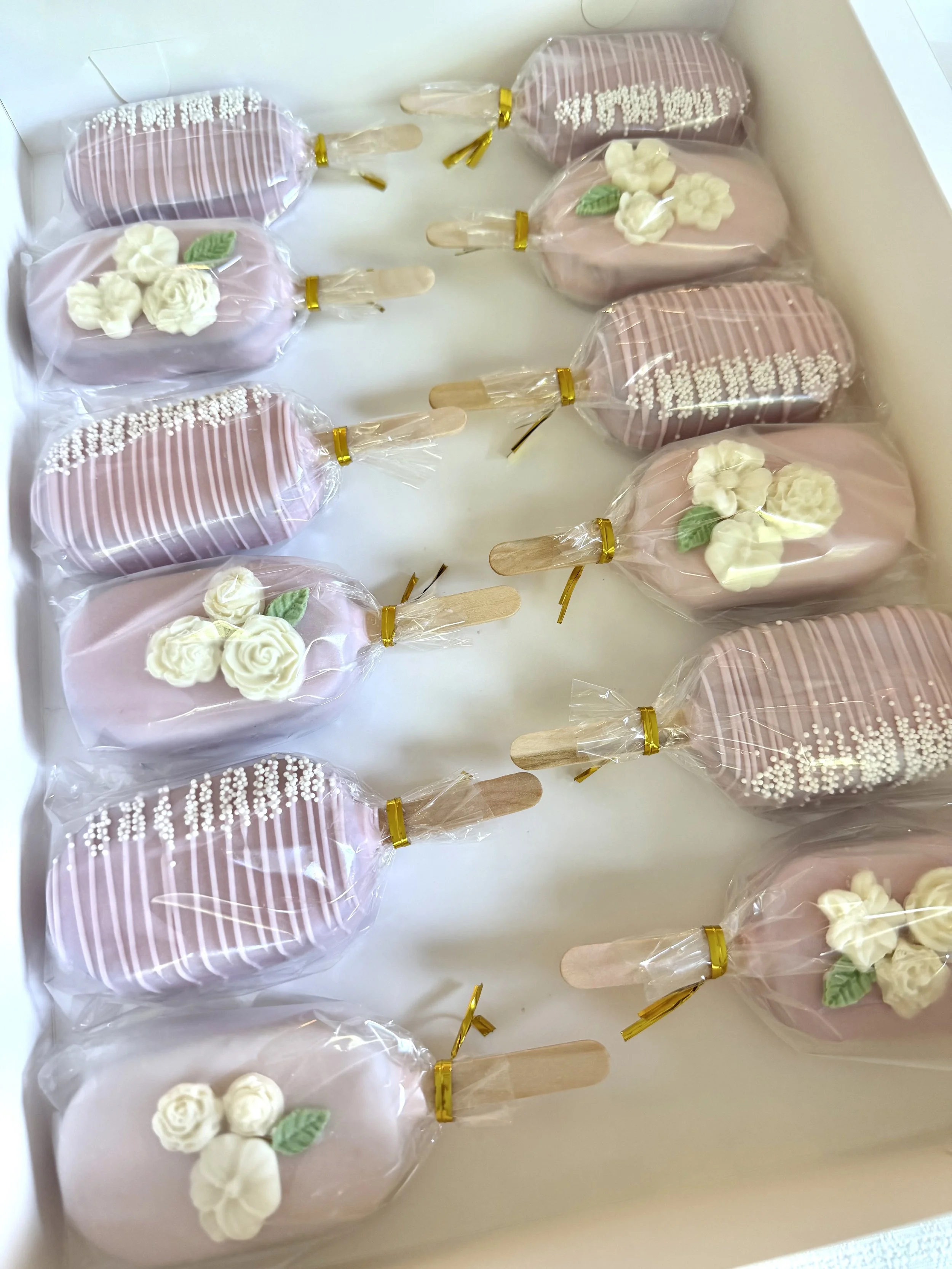 A dozen of pink cakesicles with white sprinkles and white floral decorations, individually wrapped in plastic, placed inside a white box.