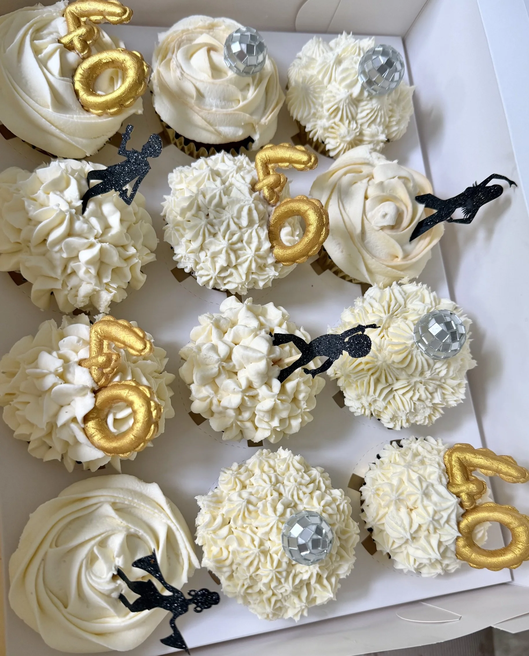 Assorted white cupcakes decorated with chocolate 40 in gold; black toppers, silver disco balls, and various cream cheese floral piping patterns, arranged in a premium white box.
