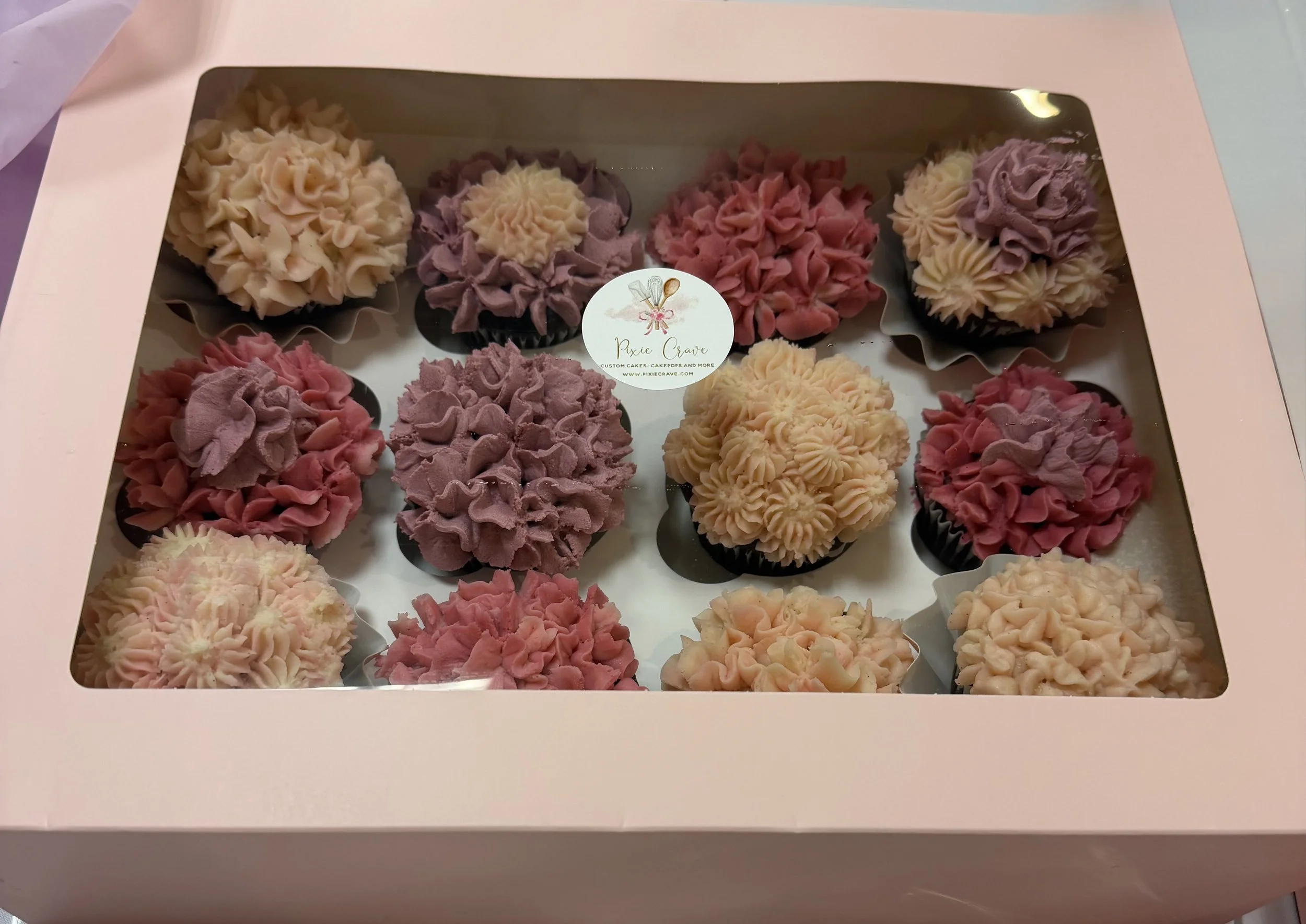 Box of colorful buttercream cupcakes with pink, purple, and cream-colored frosting.