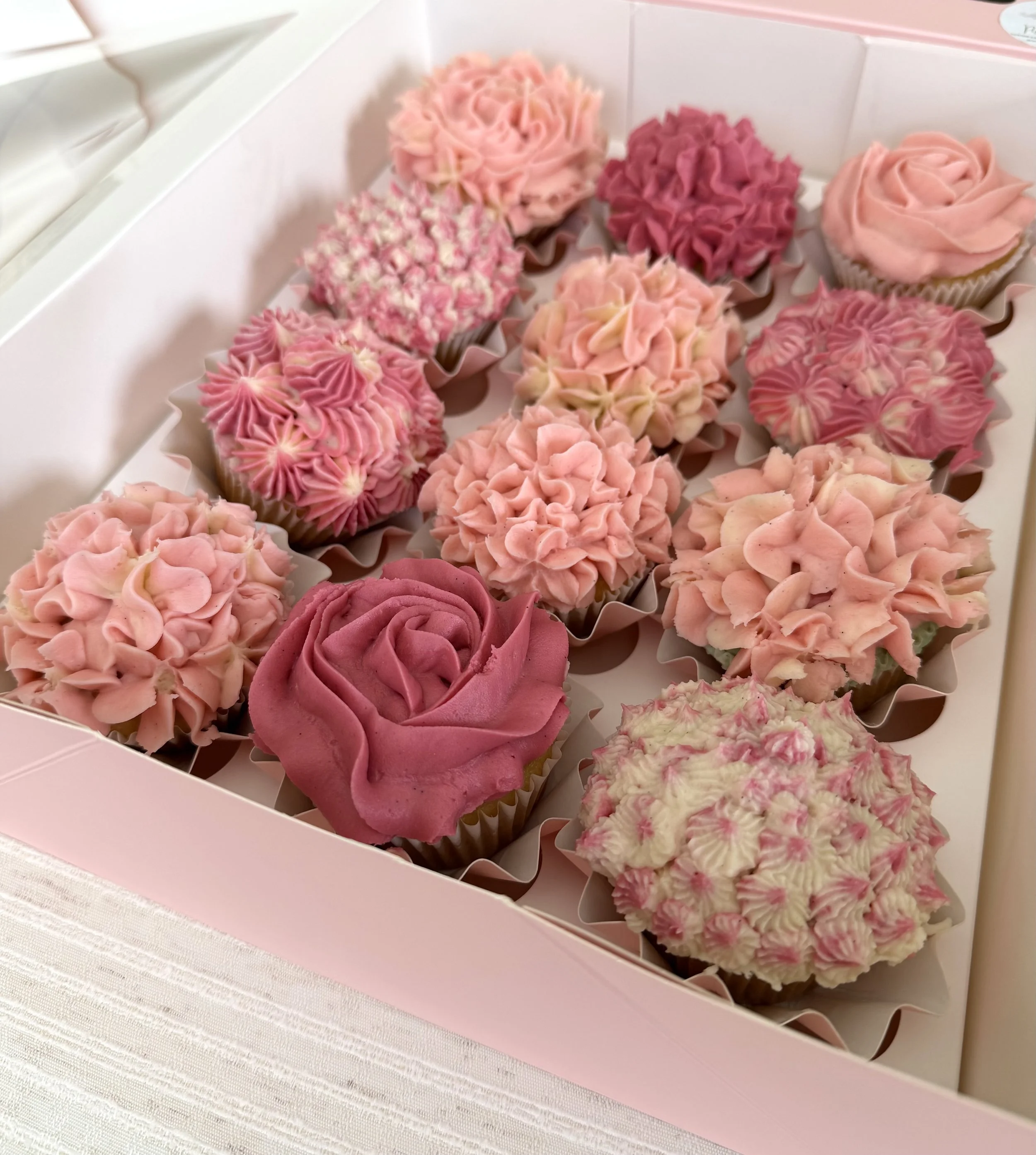 A pink cake box with twelve cupcakes decorated with pink, white, and purple icing in floral and swirling designs.