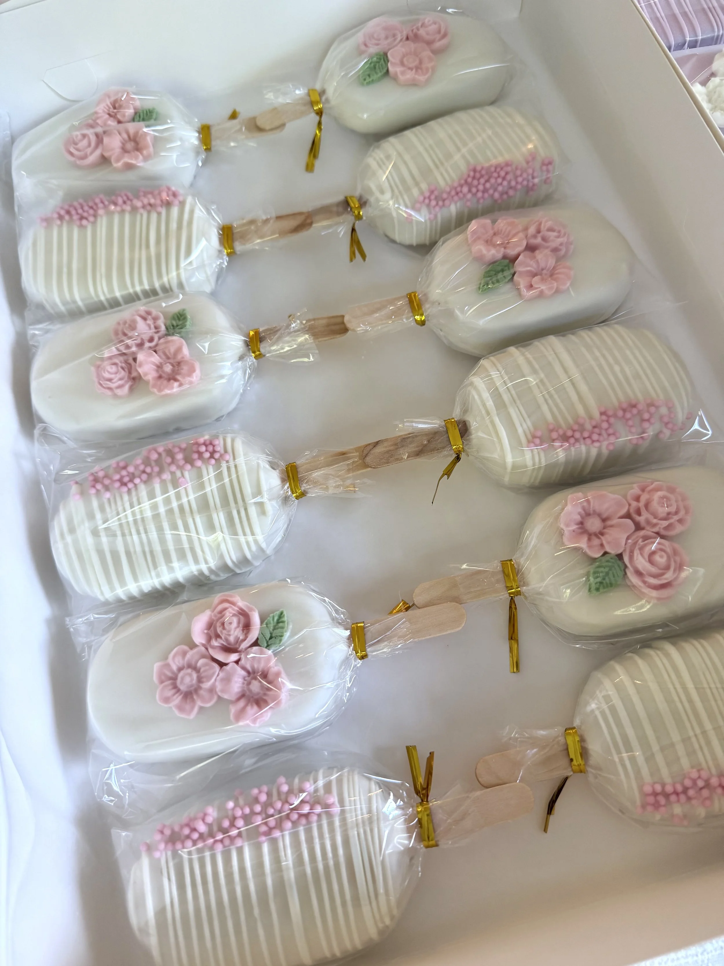 A dozen of white chocolate covered cakesicles decorated with pink and green floral chocolate, wrapped in clear plastic and tied with gold ribbons in a box.