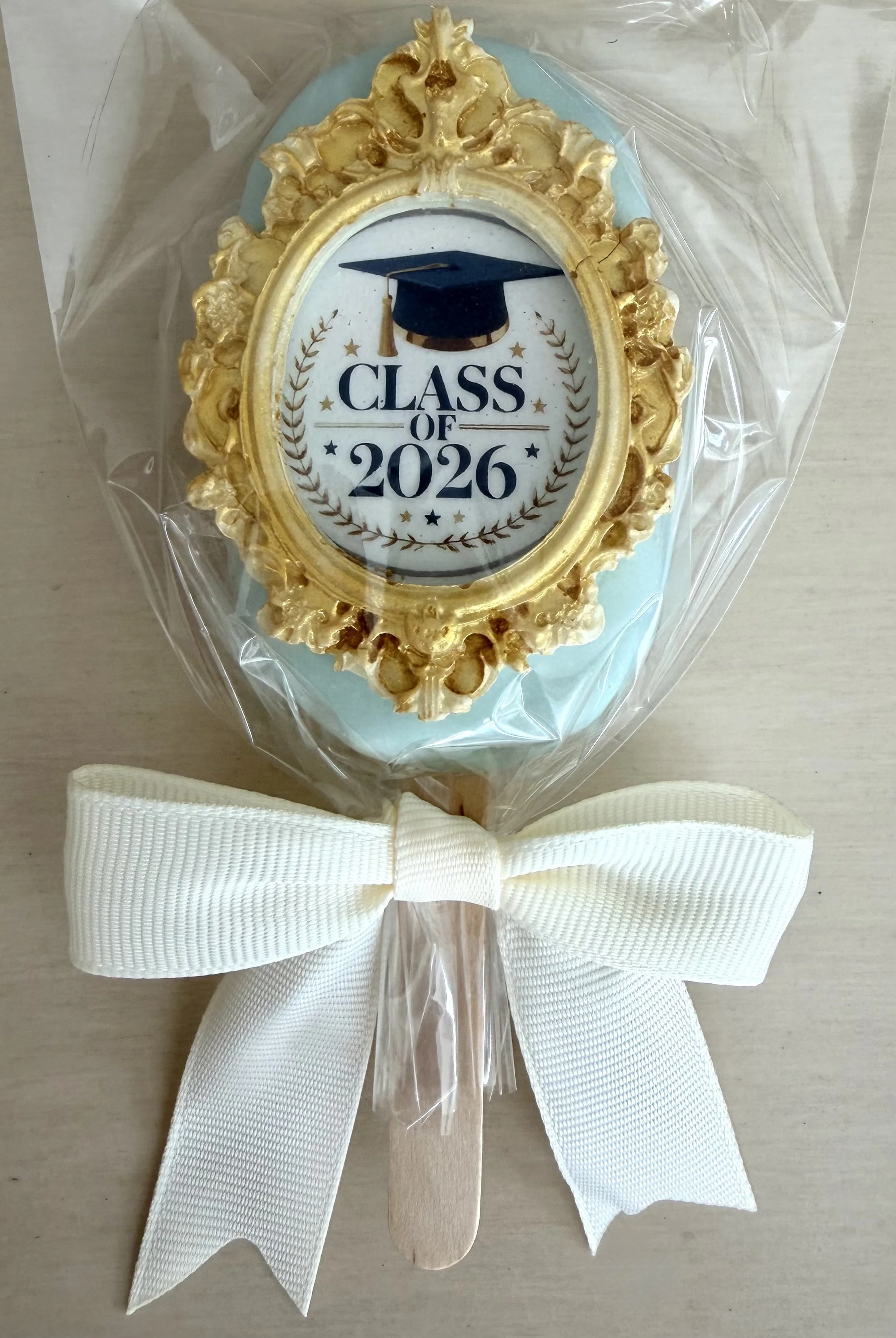 Decorative item with a gold ornate oval frame, a graduation cap and the text 'Class of 2026', wrapped in clear plastic with a white ribbon bow.