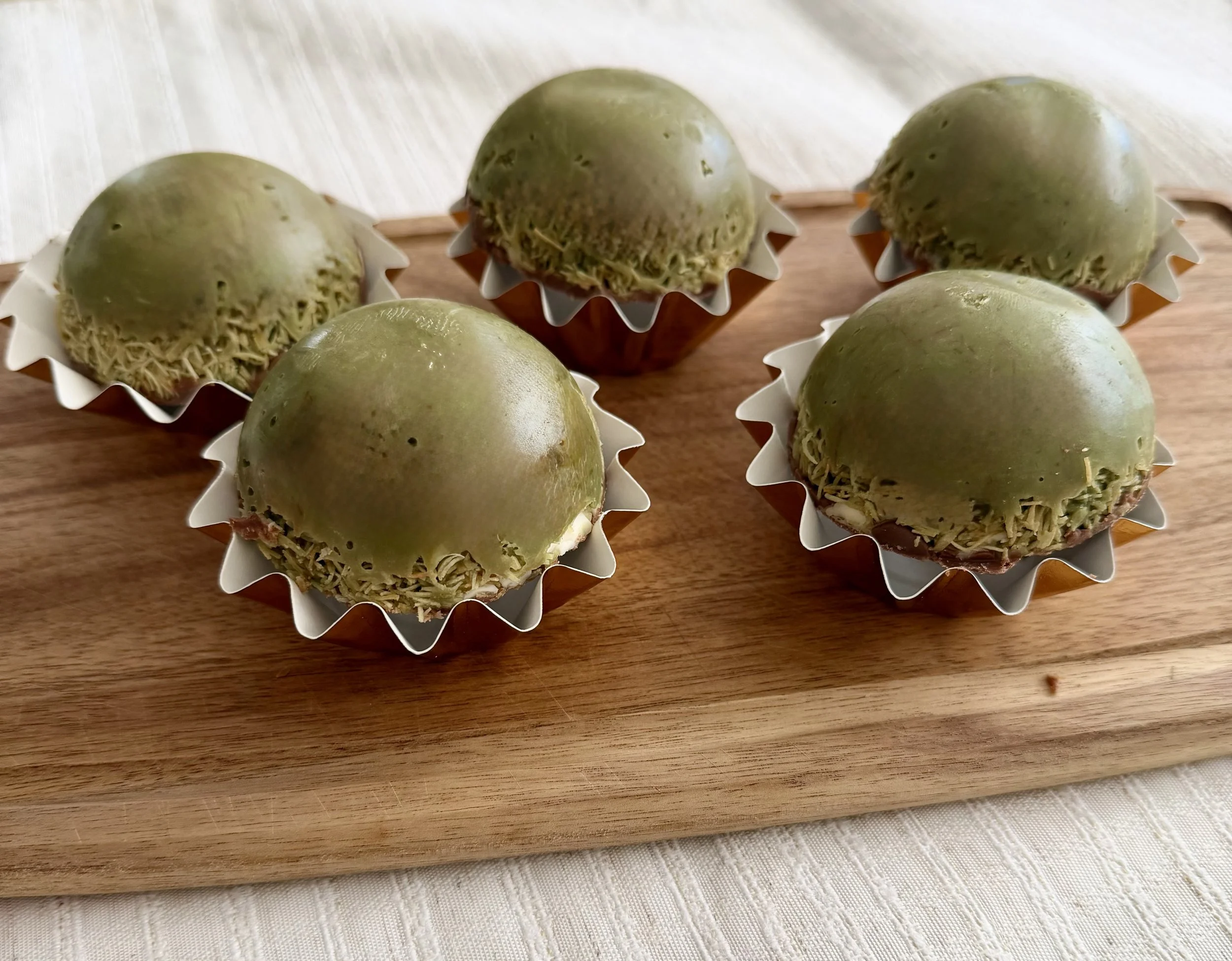 Six green pastel-colored confections with shredded coconut topping in white paper cups on a wooden tray.