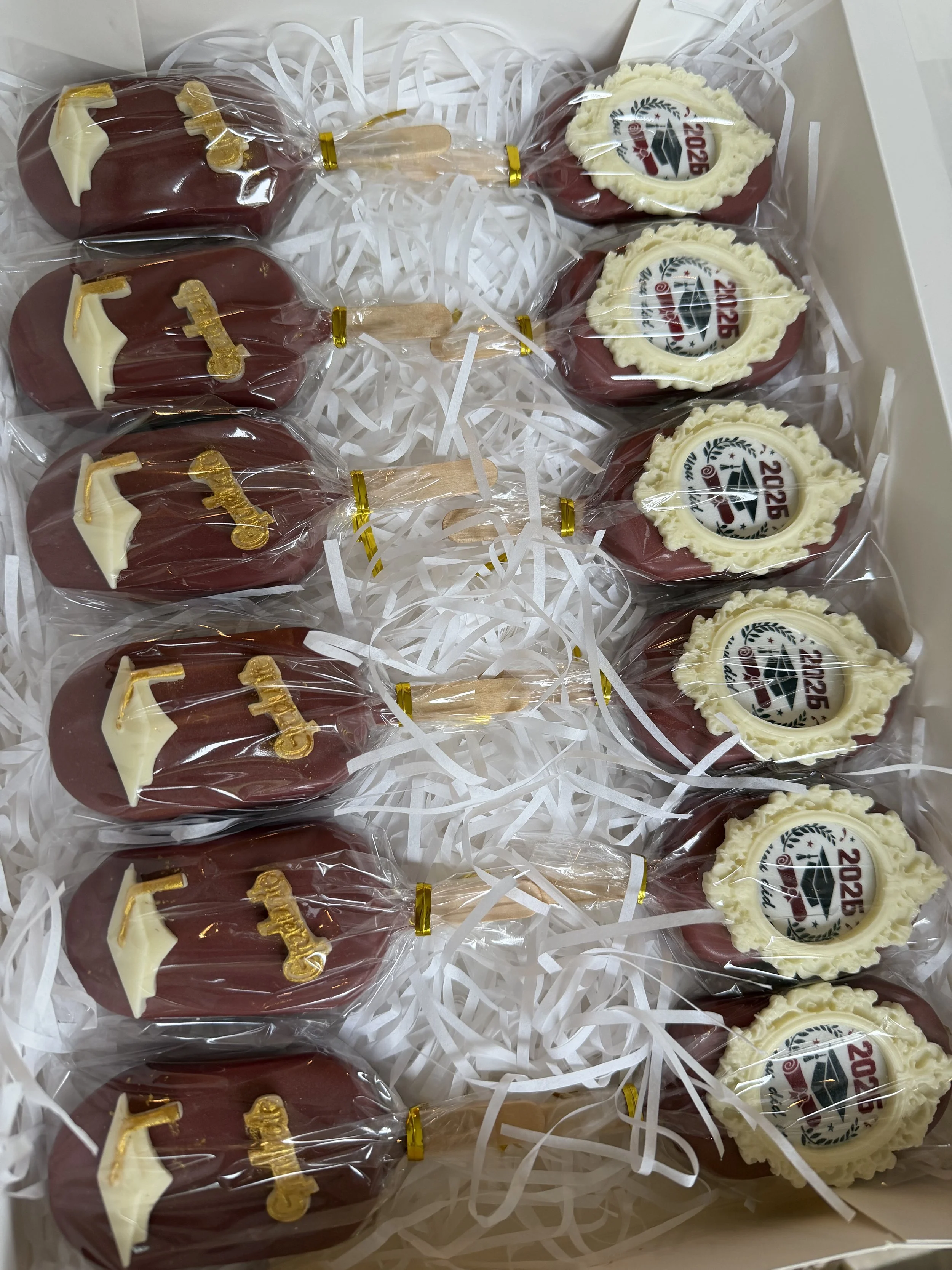 A box containing twelve decorated cakesicles wrapped in plastic. The cakesicles have a dark red and white color graduation theme with football decor and the year 2026.