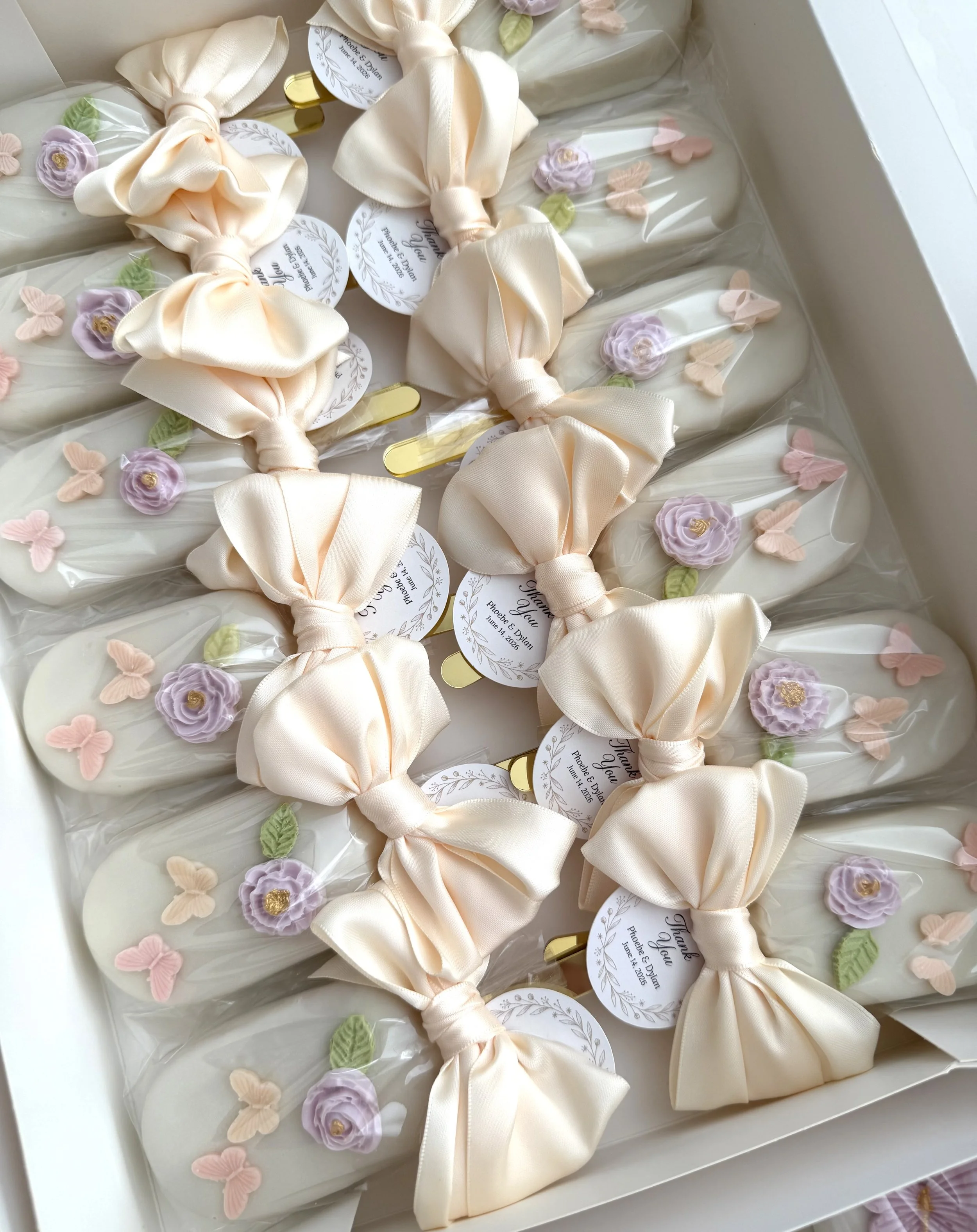 A dozen of decorated cakesicles wrapped with off-white satin bows and pastel floral accents in purple, pink, and green. Golden sticks and personalized tags for a wedding celebration.