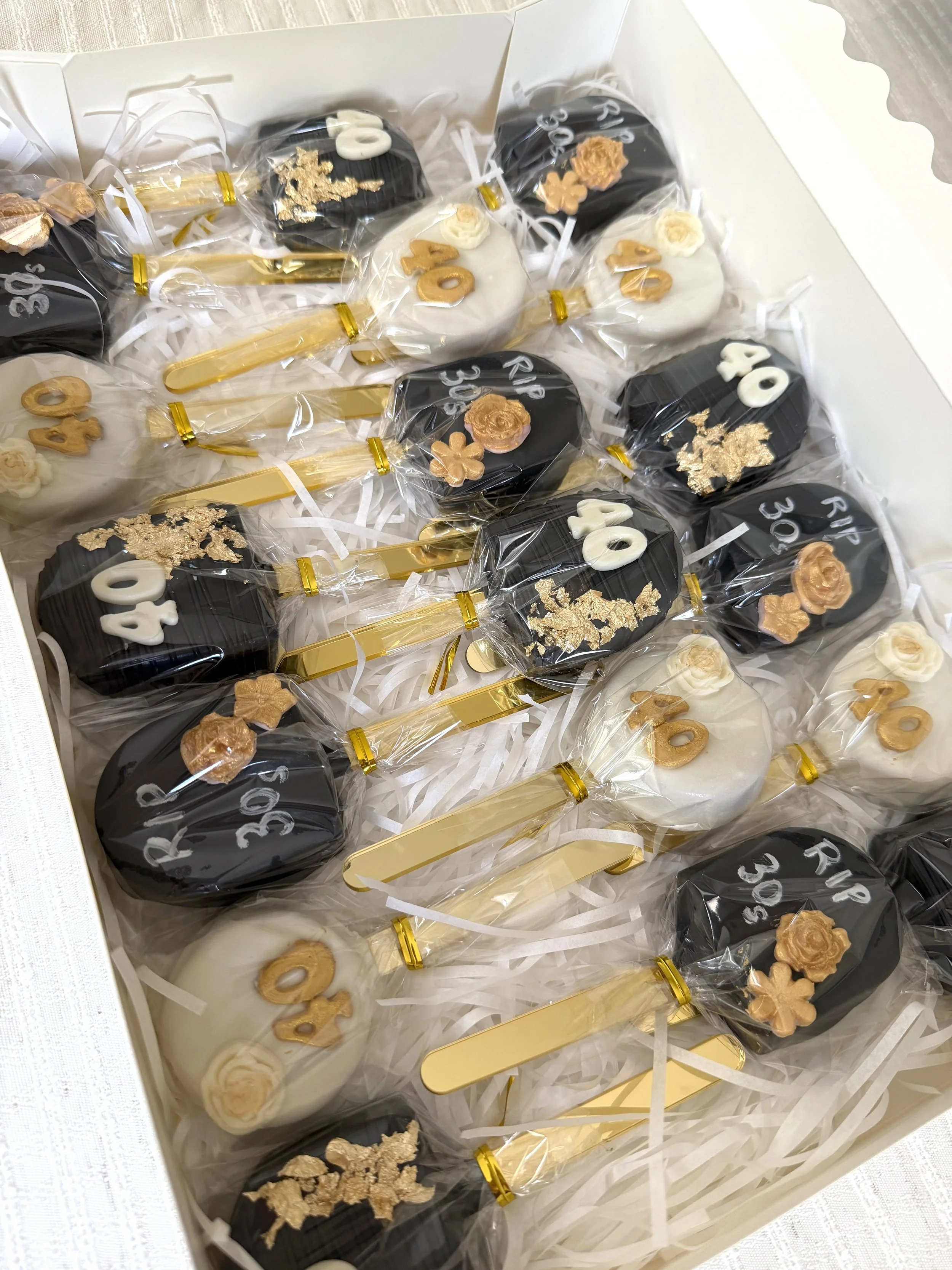 A white gift box filled with assorted decorated cakepops wrapped in clear plastic with gold ribbons, including black and white 40th Birthday theme with gold accents.