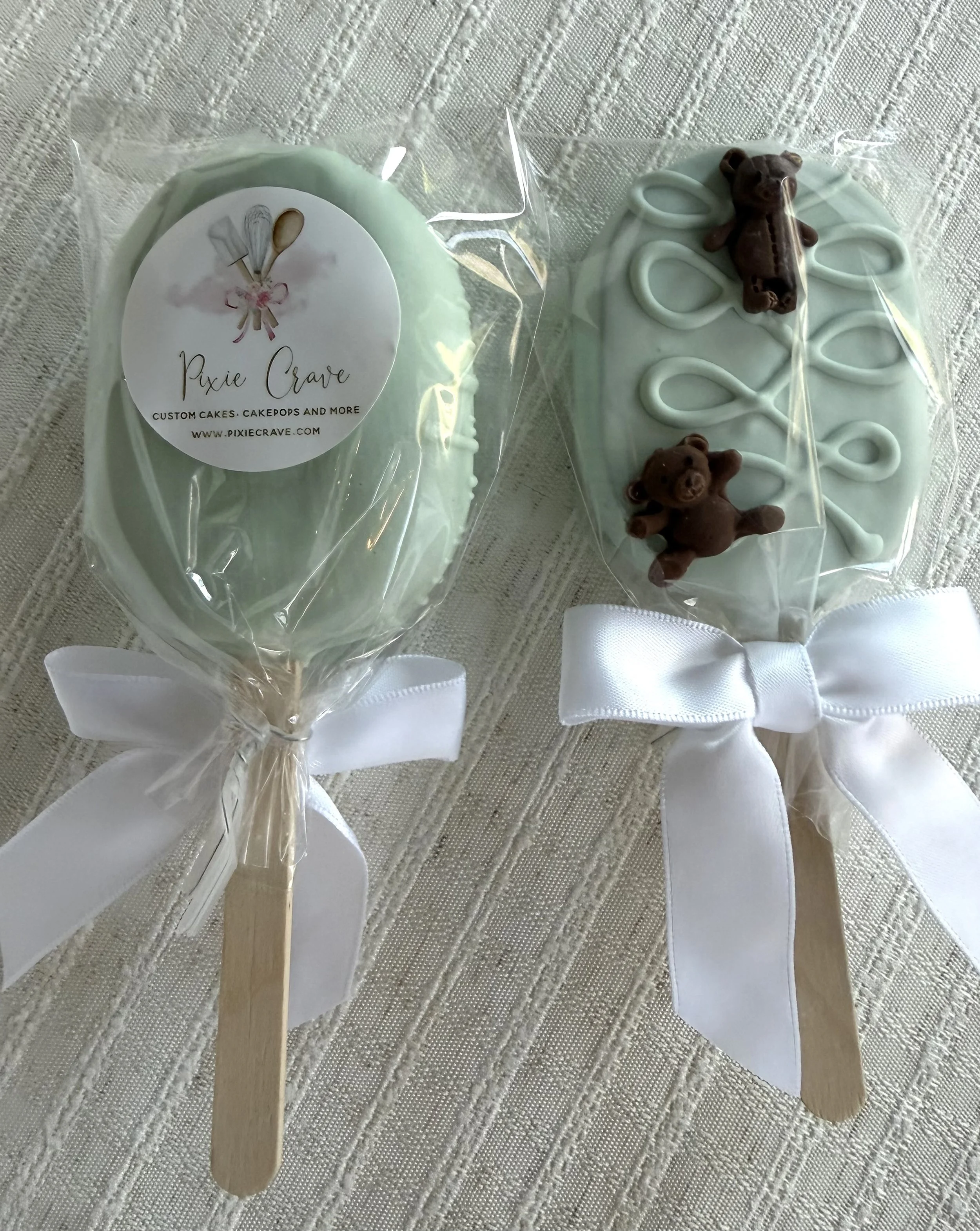 Decorative soap or craft item shaped like a popsicle with a white ribbon and a teddy bear figure on top, wrapped in clear plastic with a white ribbon at the bottom, placed on a textured fabric surface.