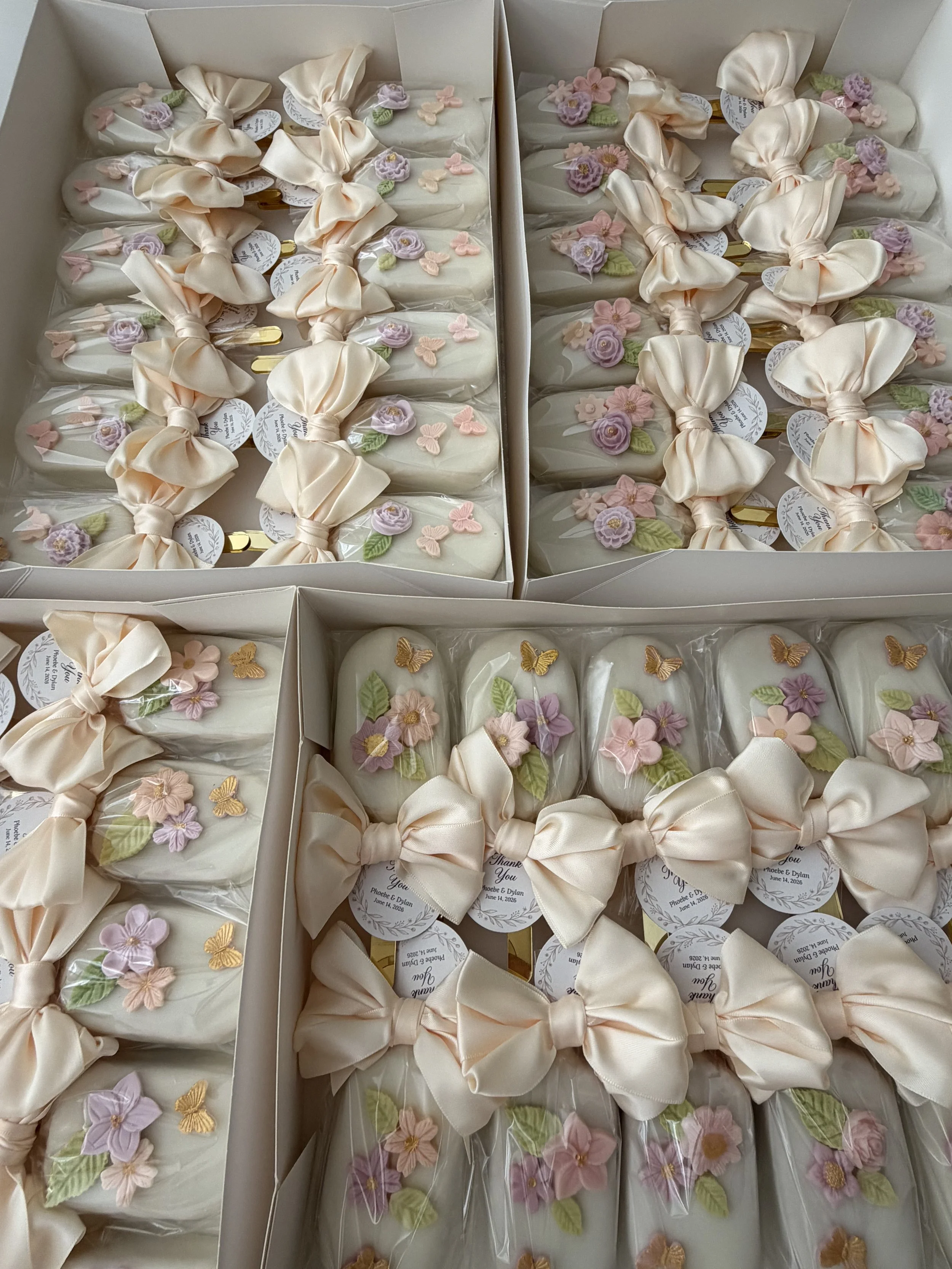 Boxes of decorative wedding favors wrapped in cream-colored satin ribbon, adorned with purple, pink, and pastel-colored floral and butterfly embellishments, with tags that read 'Thank You' and details of a wedding date.