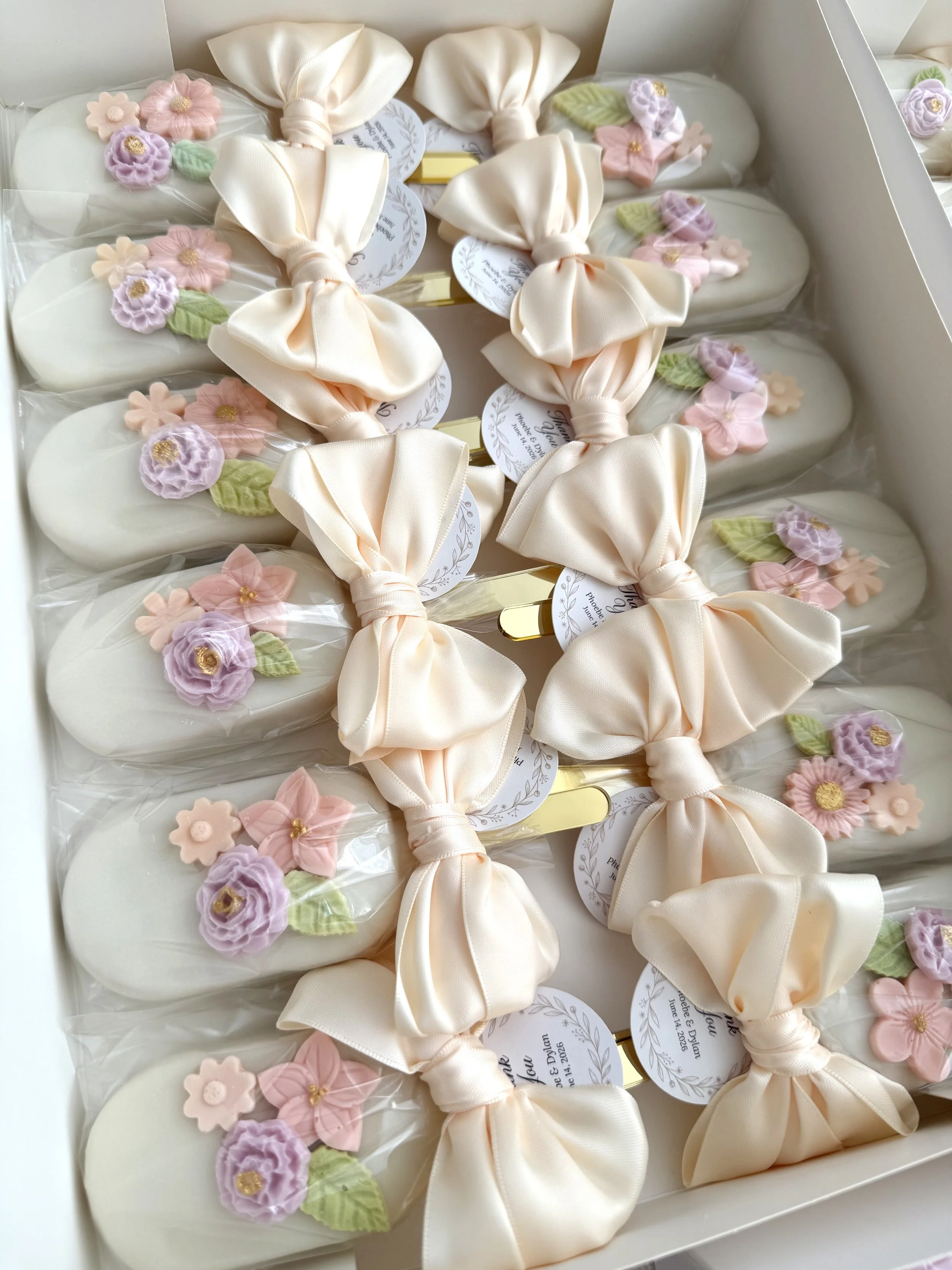 A dozen of decorated cakesicles wrapped with off-white satin bows and pastel floral accents in purple, pink, and green. Golden sticks and personalized tags. Perfect for wedding favors!
