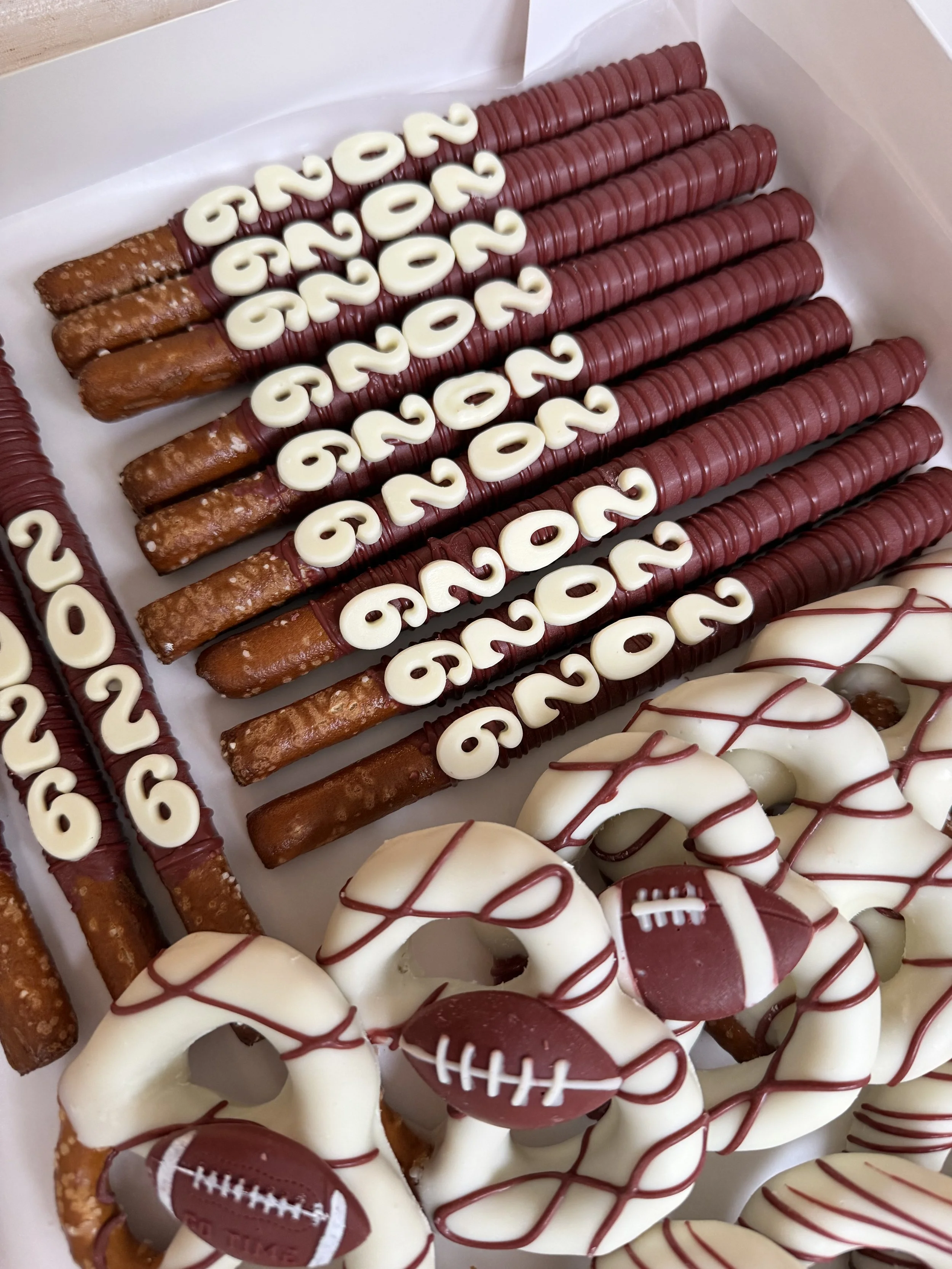 A box of decorated dipped pretzels with football theme. The tall and round pretzels have footballs, graduation Class of 2026 rings, and other sports symbols, decorated with white and red chocolate and drizzles.