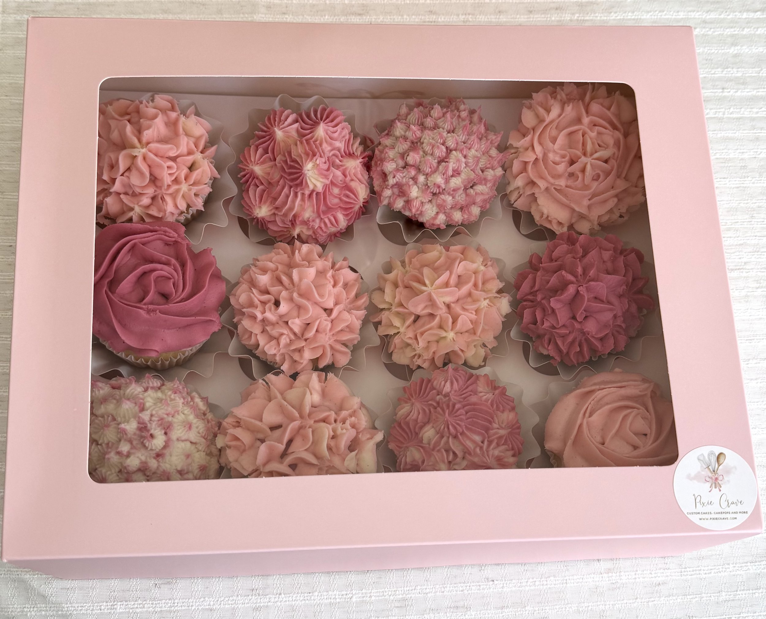 Pink box with a clear window displaying an assortment of pink and white frosted cupcakes with various decorative styles, placed on a white cloth surface.