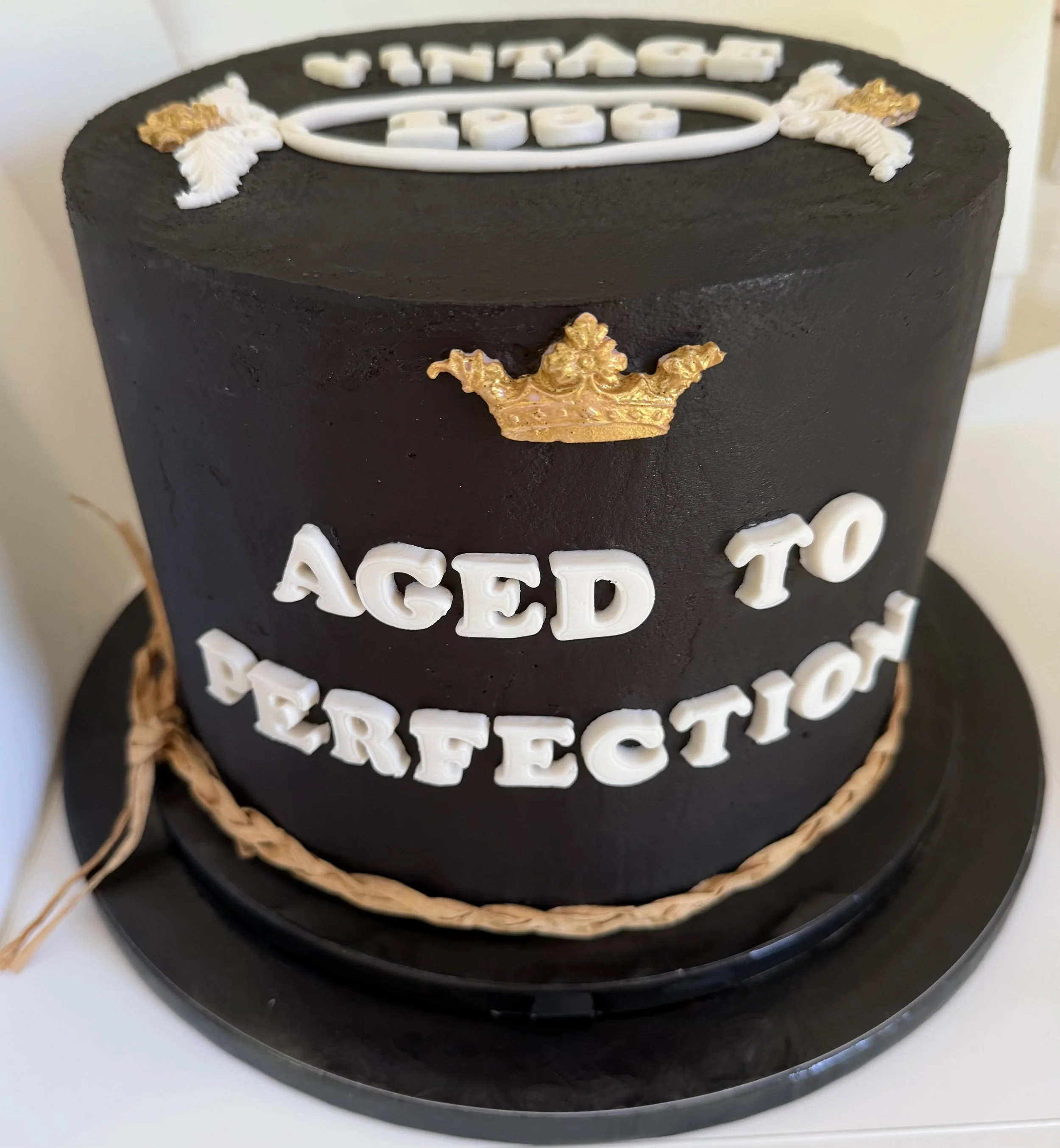 A black birthday cake with white fondant letters spelling out "Vintage 1986" on top and a golden crown decoration on the side with fondant letters "aged to perfection". The cake is wrapped with a decorative brown raffia ribbon at the base.