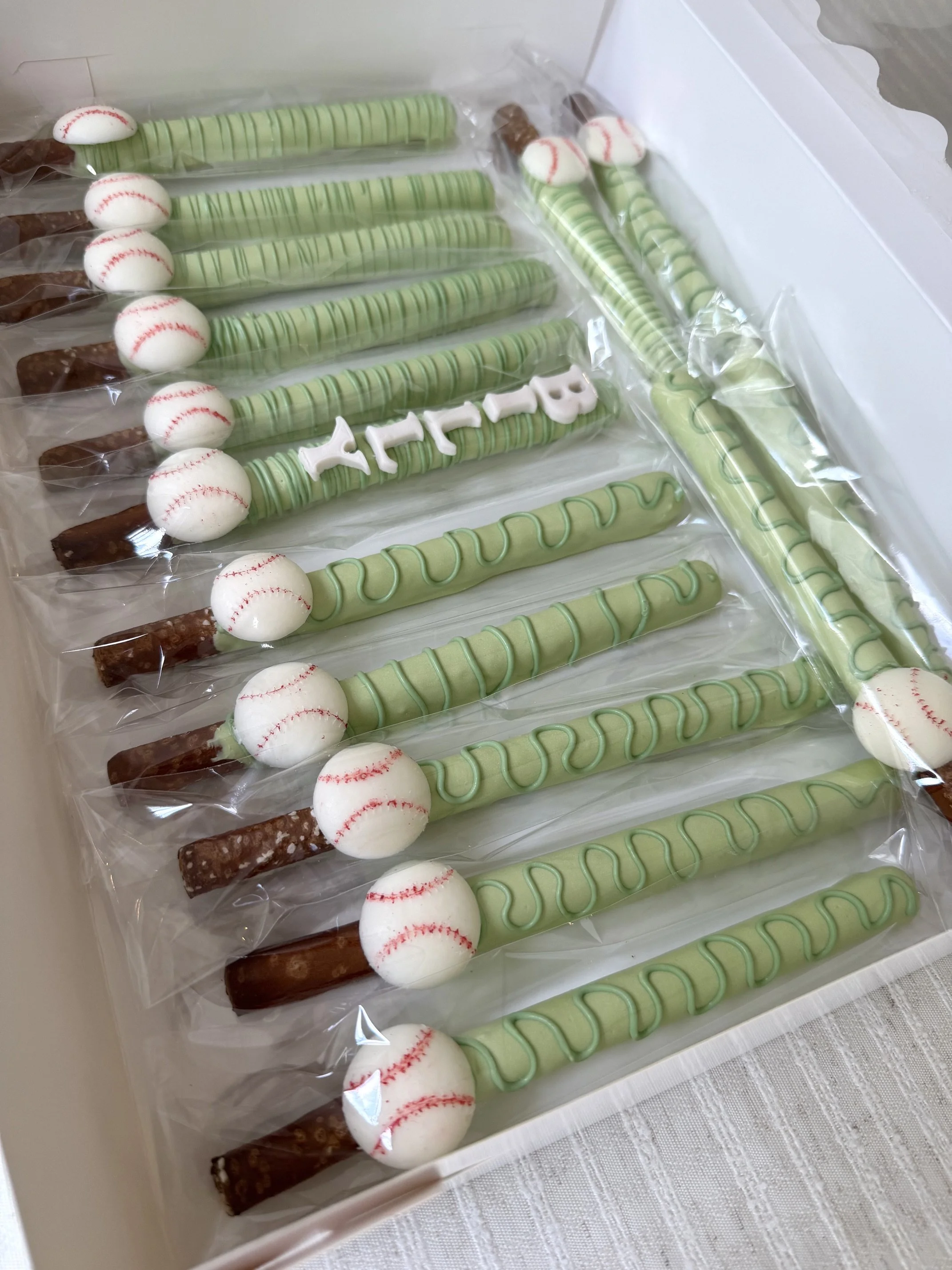 Box of baseball-themed dipped pretzels with white spherical tops and red stitches, decorated with green and white striped chocolate melt.