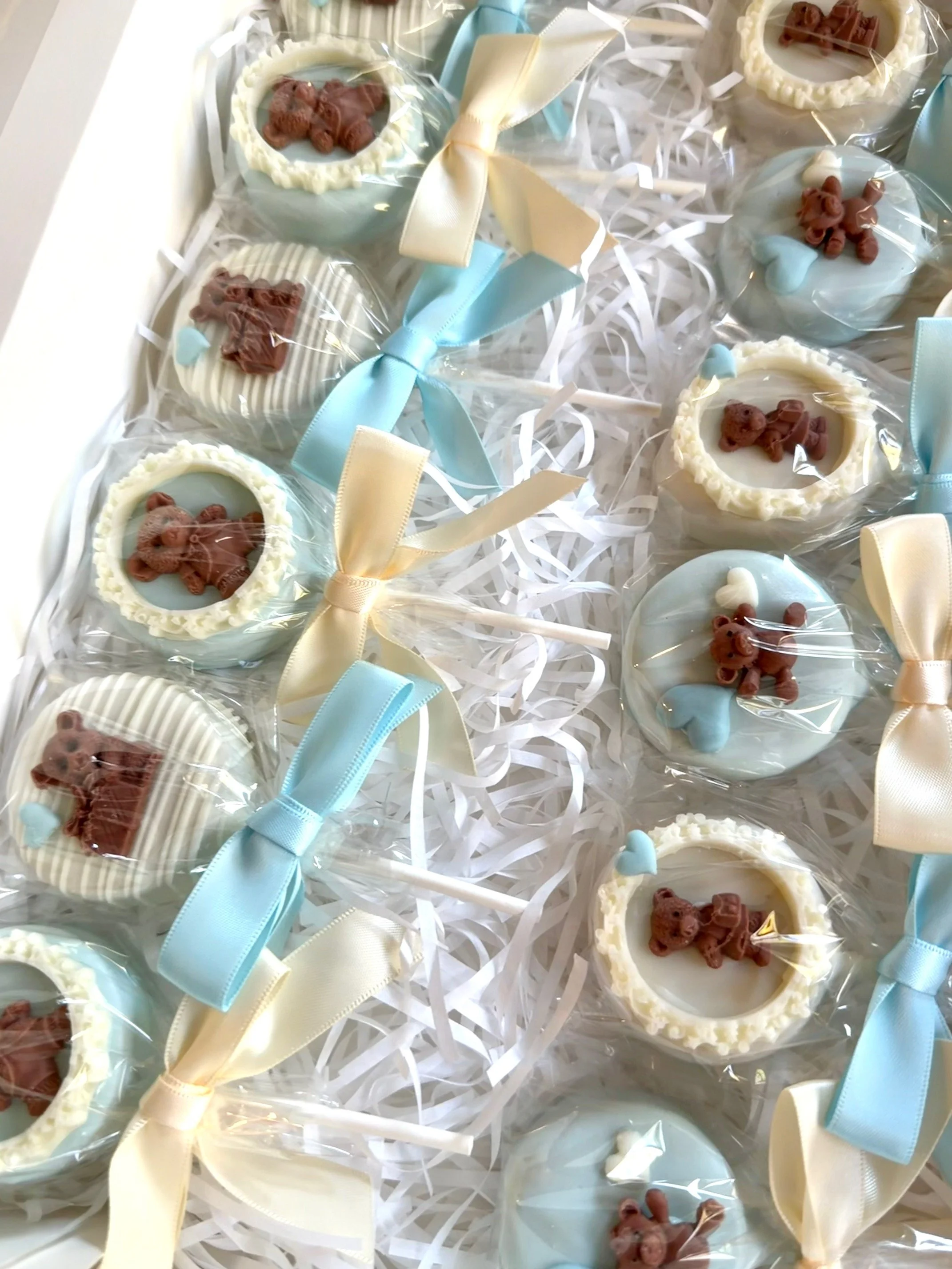 A dozen of decorated chocolate teddy bear themed cake pops wrapped in clear plastic with pastel-colored ribbons baby blue and ivory for a special baby shower.