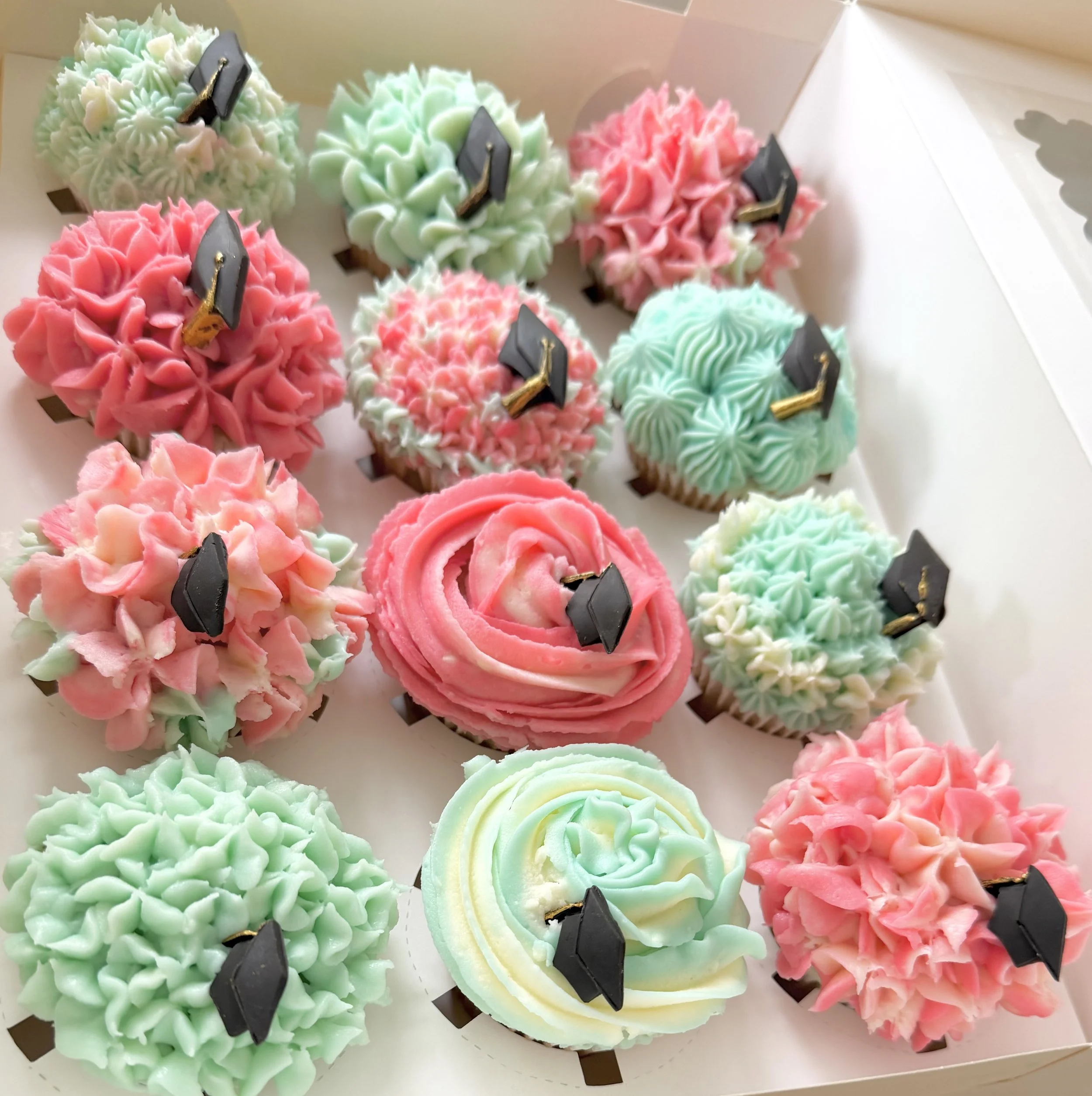 Colorful cupcakes decorated with pink, green, and white frosting, each topped with a small black chalkboard sign with gold holder in a white box.